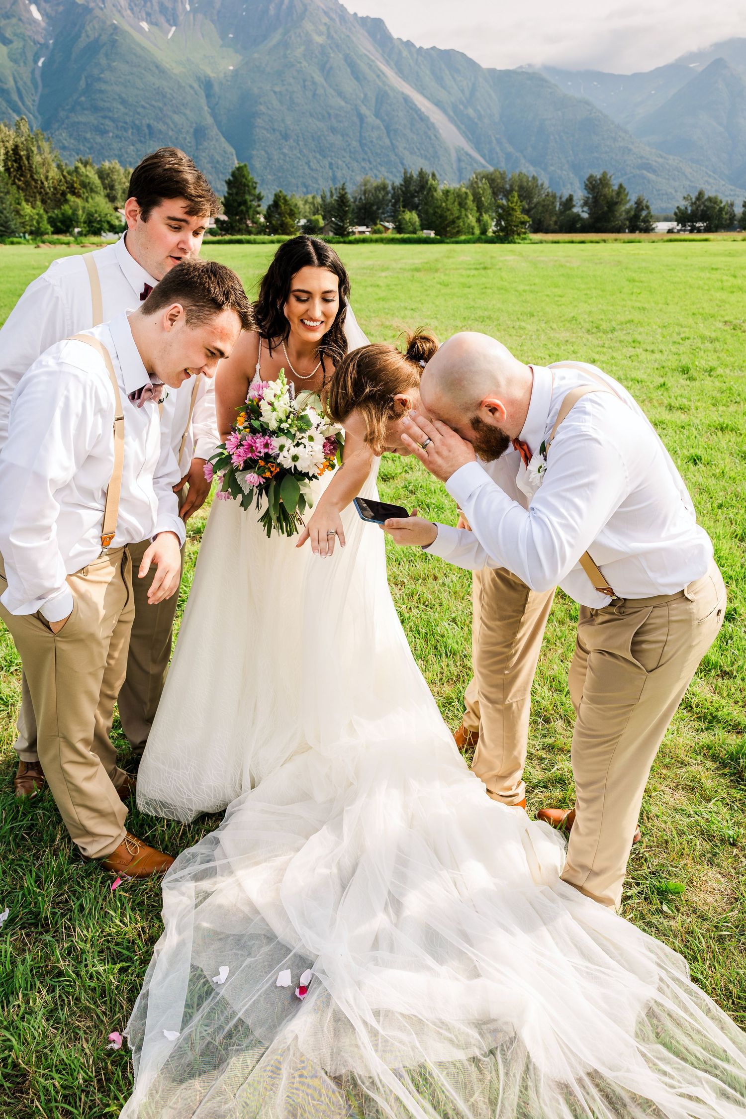 Wedding party gathers around bride's flowing veil in front of majestic mountain landscape.