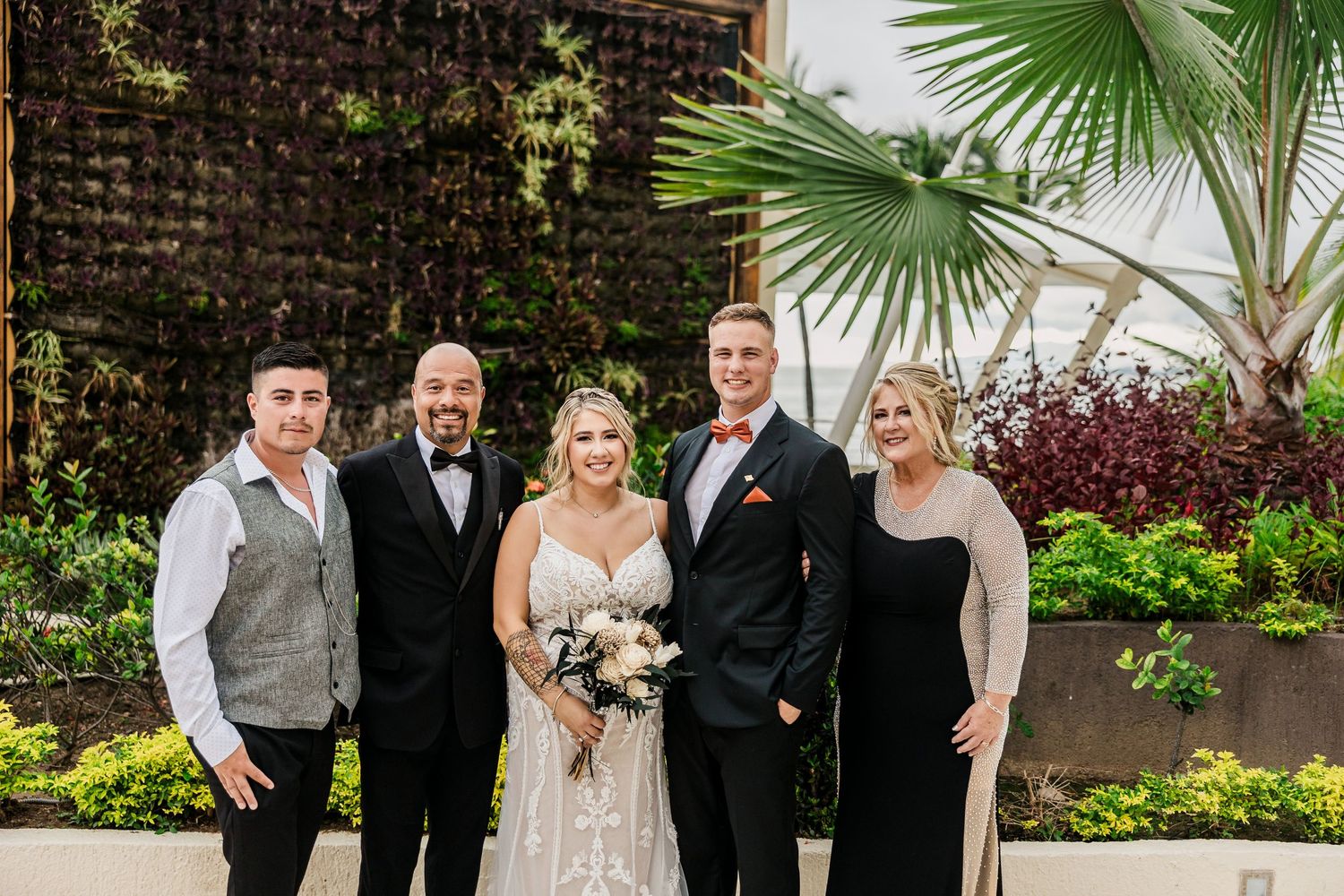 Wedding party posing outdoors with lush greenery, bride in white gown holding bouquet, groom in black tuxedo.