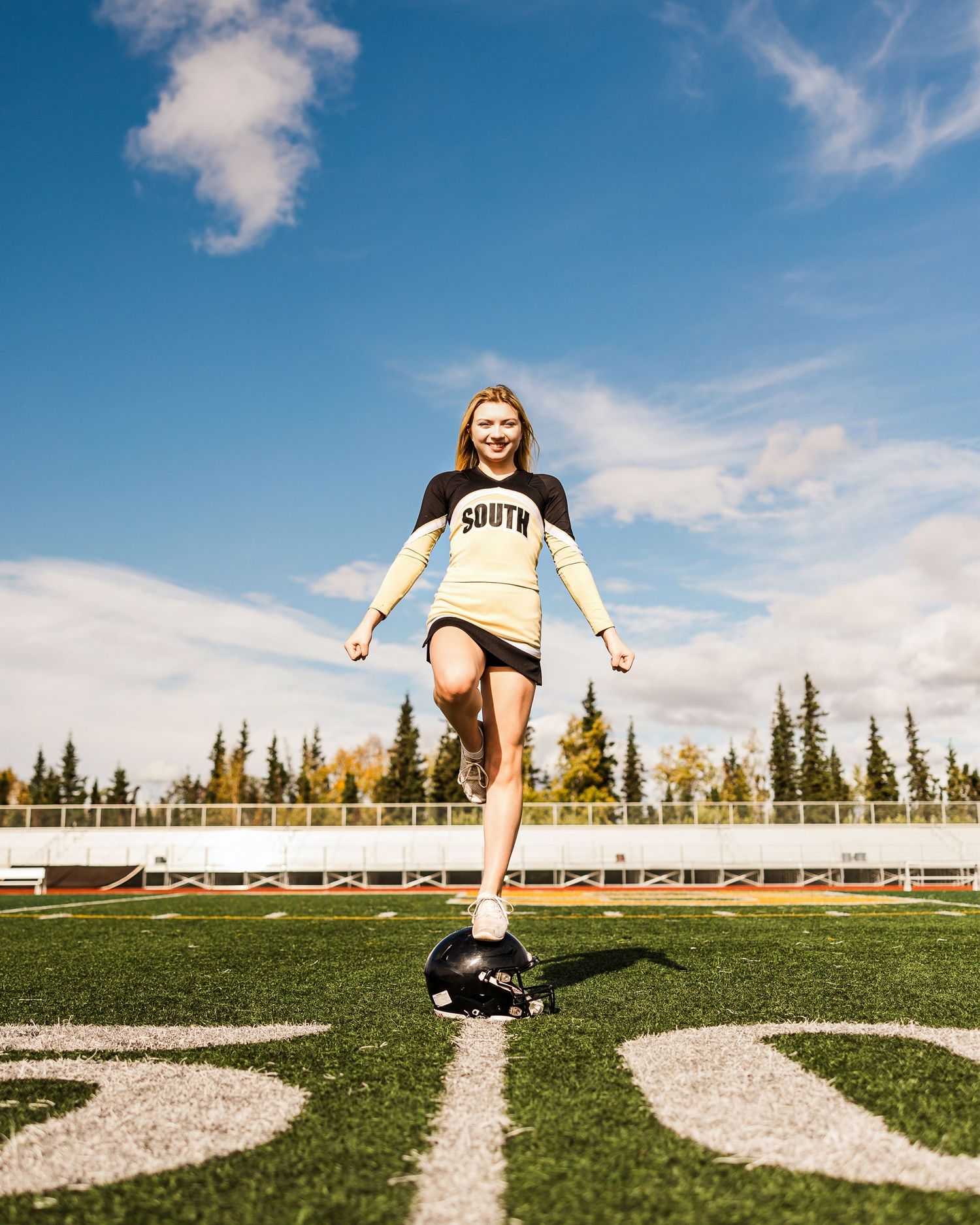 Young woman in a South jersey steps on a helmet on a sunny football field with trees behind.