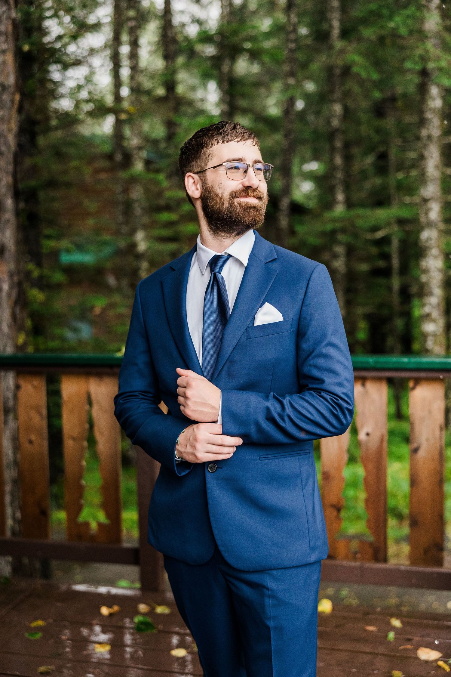 A person in a navy suit and glasses poses on a wooden deck in a forest setting.