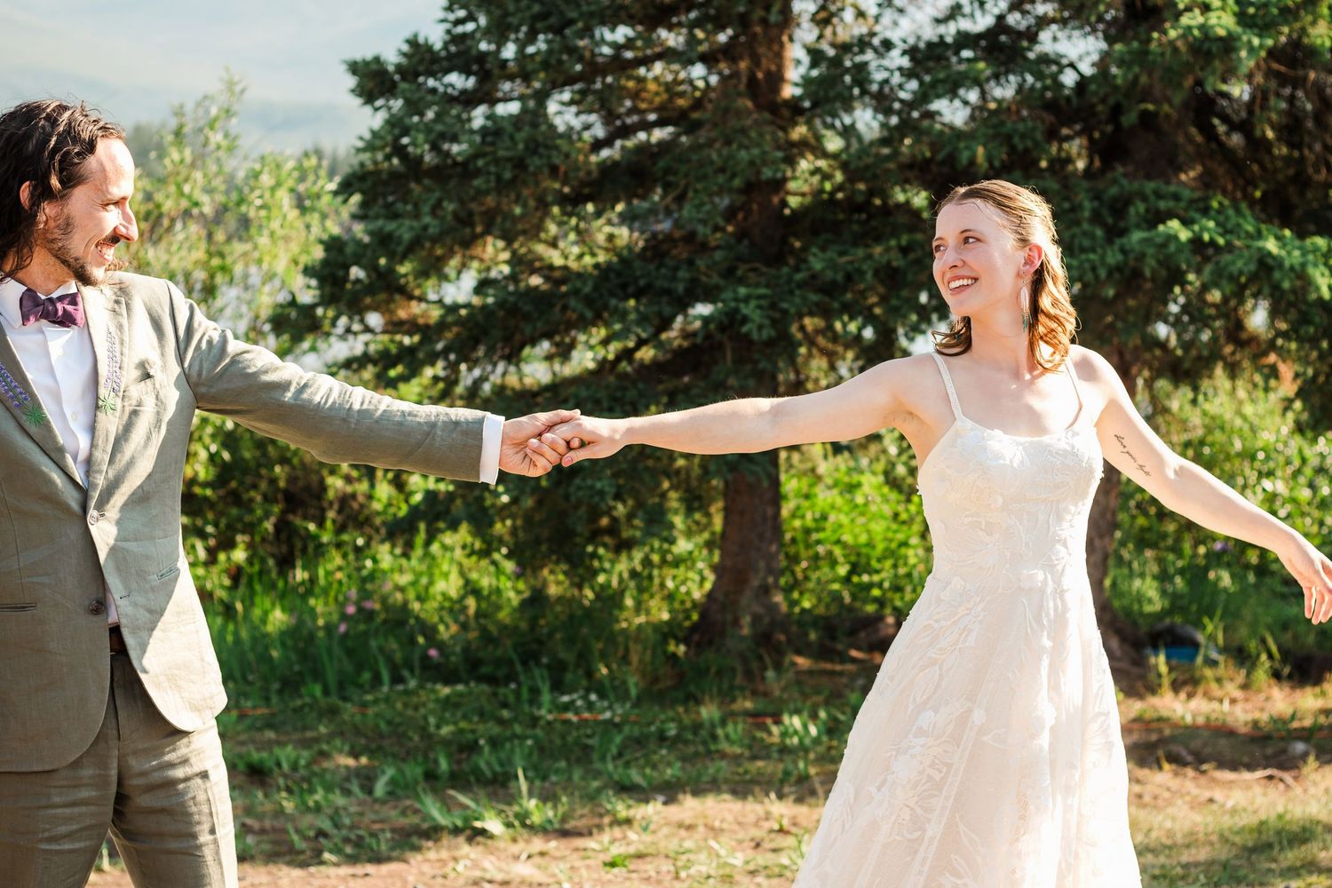 A couple in wedding attire holds hands while posing in a sunlit outdoor setting with evergreen trees.