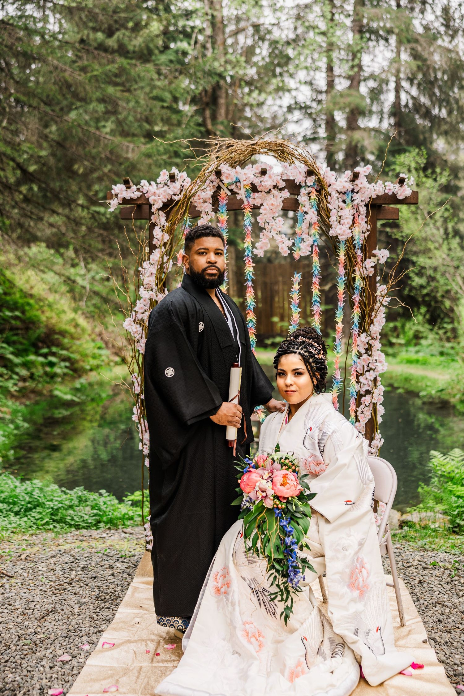 Couple in traditional Japanese wedding attire posing under a floral sakura arch in forest.