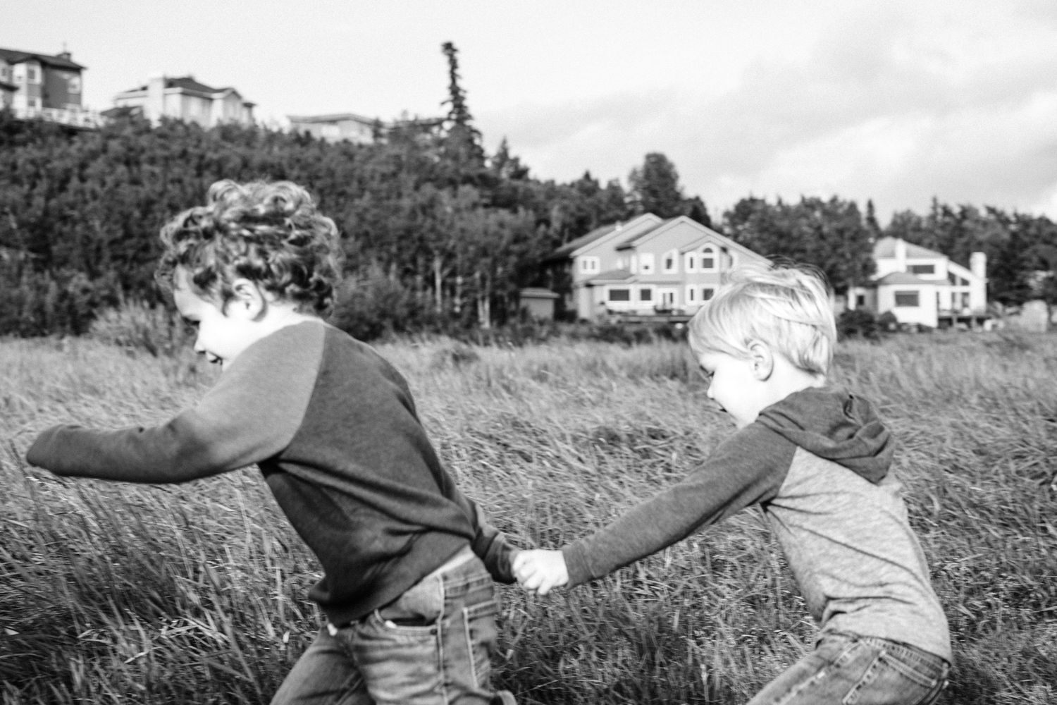 Two children play in tall grass in a black and white photo with houses and trees in background.
