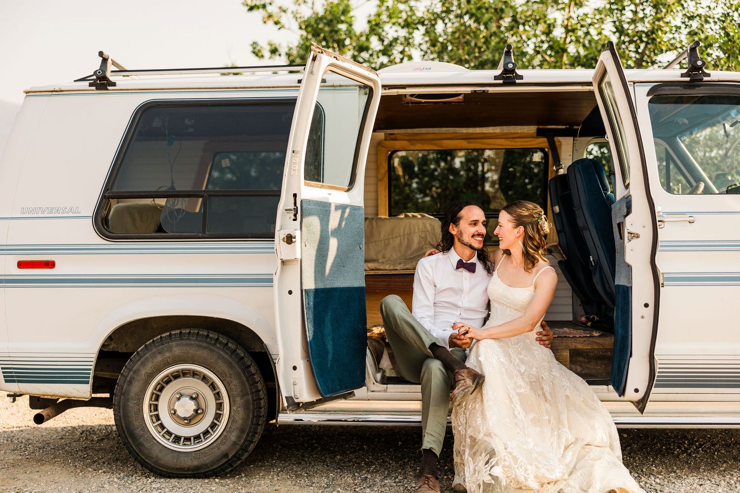 A couple in wedding attire sits in the doorway of a vintage white van.
