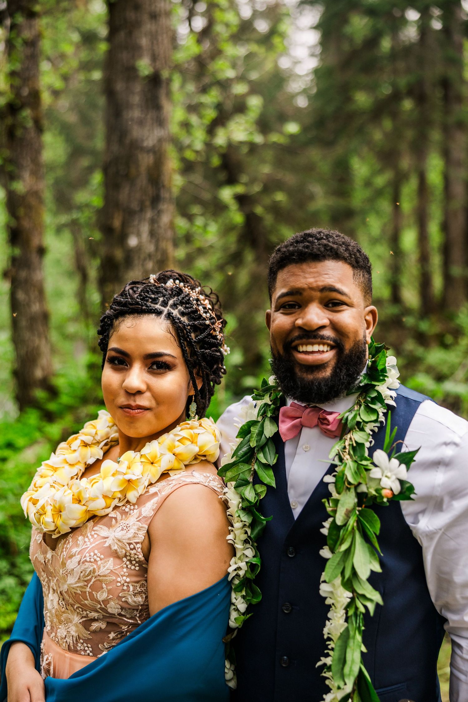 A wedding couple wearing traditional leis pose together in a forest setting.