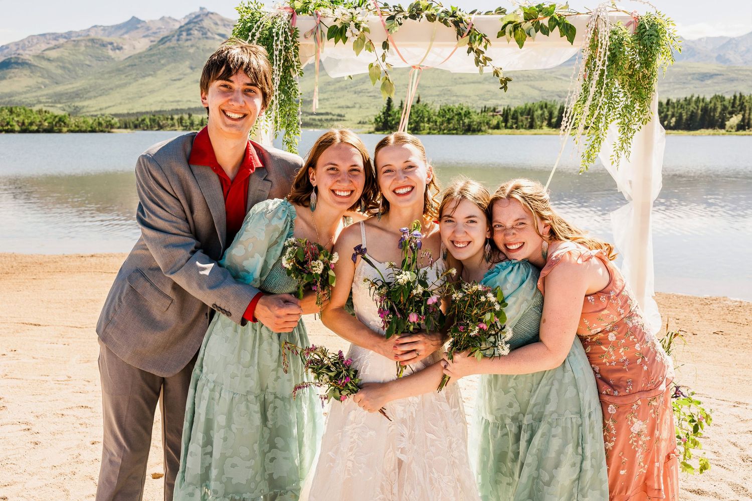 Wedding party group hug on lakeside beach with mountains in background.