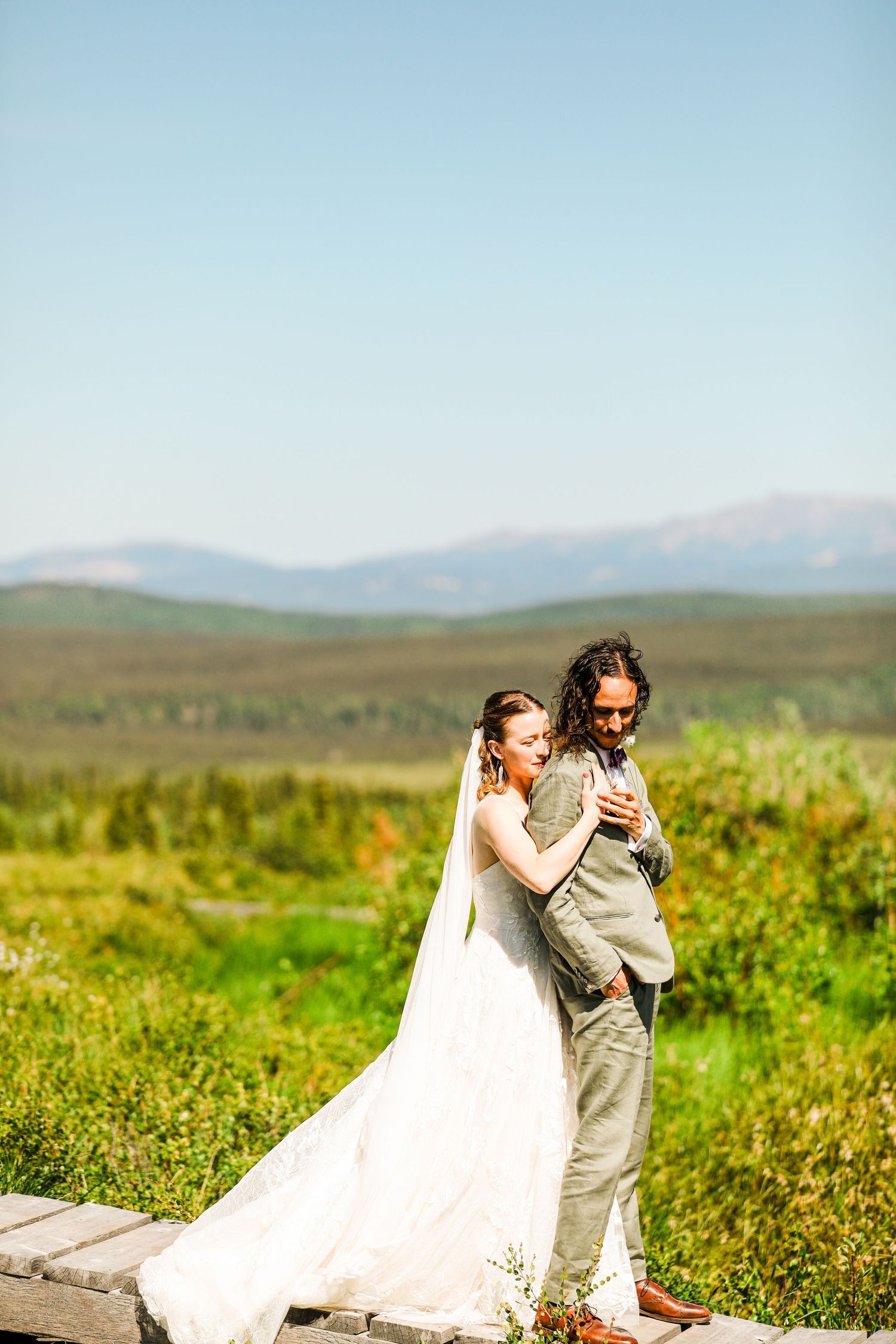 Couple embraces on wooden walkway surrounded by lush mountain landscape.