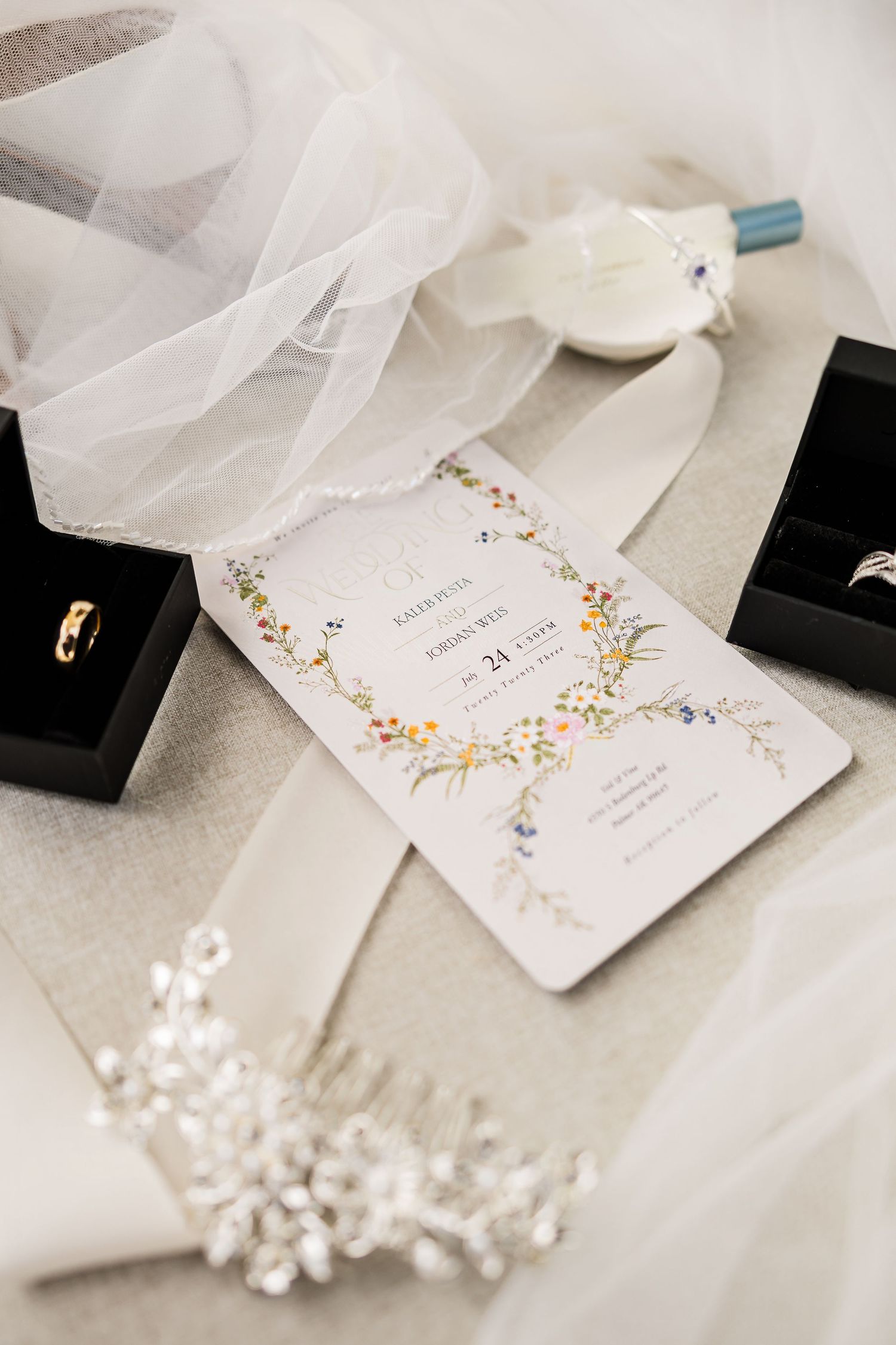 Elegant wedding flat lay with floral invitation, rings, veil, boutonniere, and crystal hair piece.