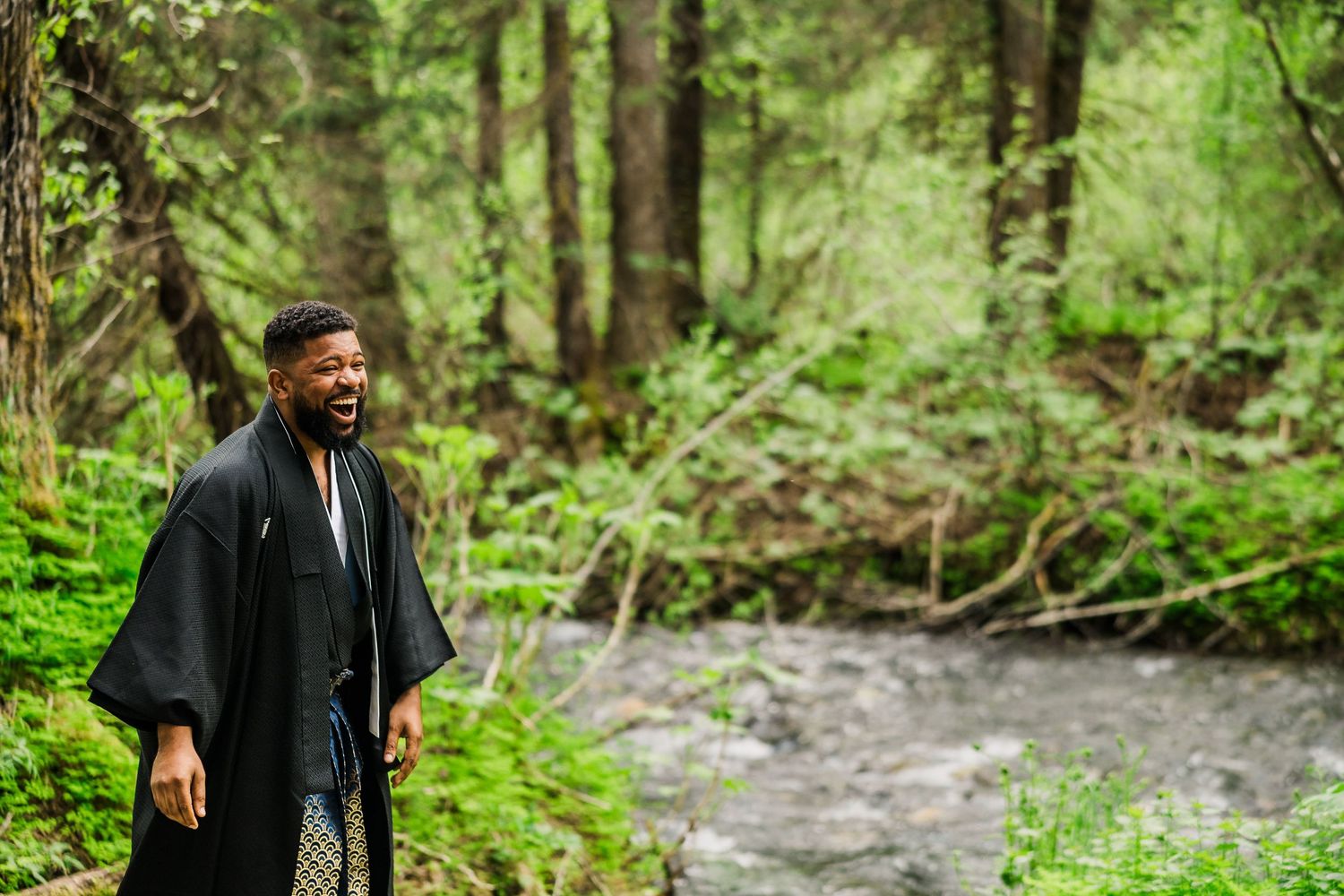 Groom in black traditional robe smiles during outdoor forest wedding ceremony.