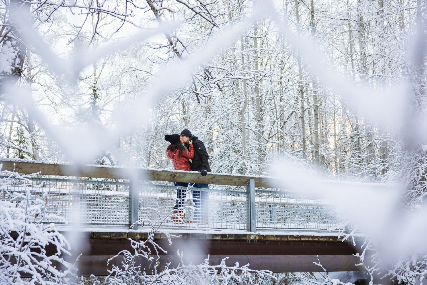 Couple kisses on a snow-covered bridge surrounded by frost-covered trees in a winter forest.