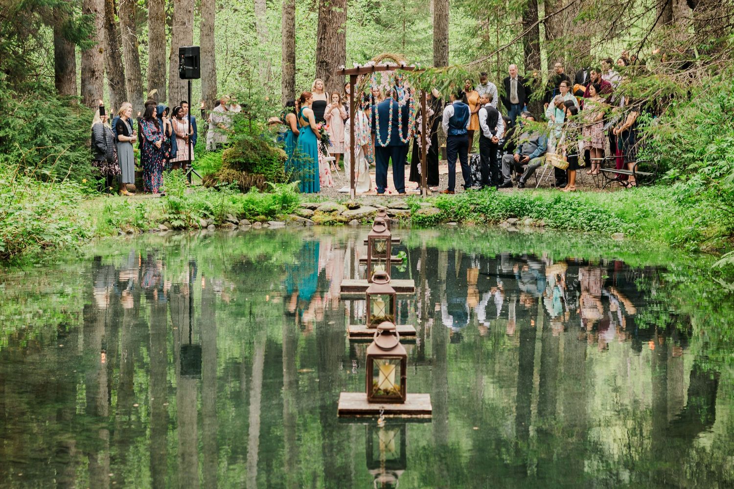 Serene pond surrounded by forest trees with guests gathered for an outdoor wedding ceremony.