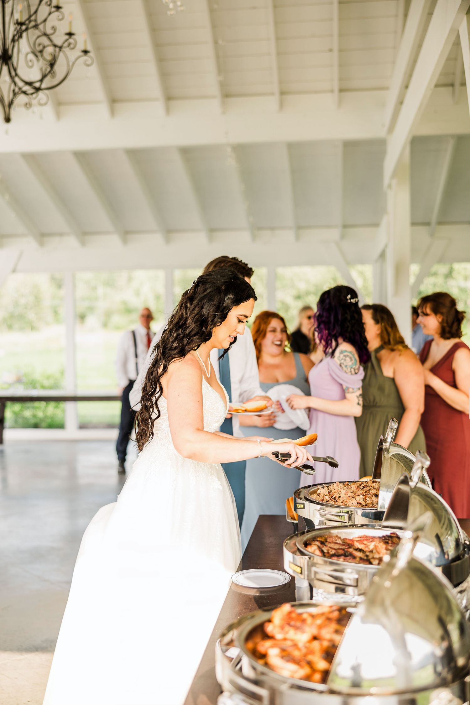 A bride serves food from a buffet line at a wedding reception while guests line up in a white-covered pavilion.
