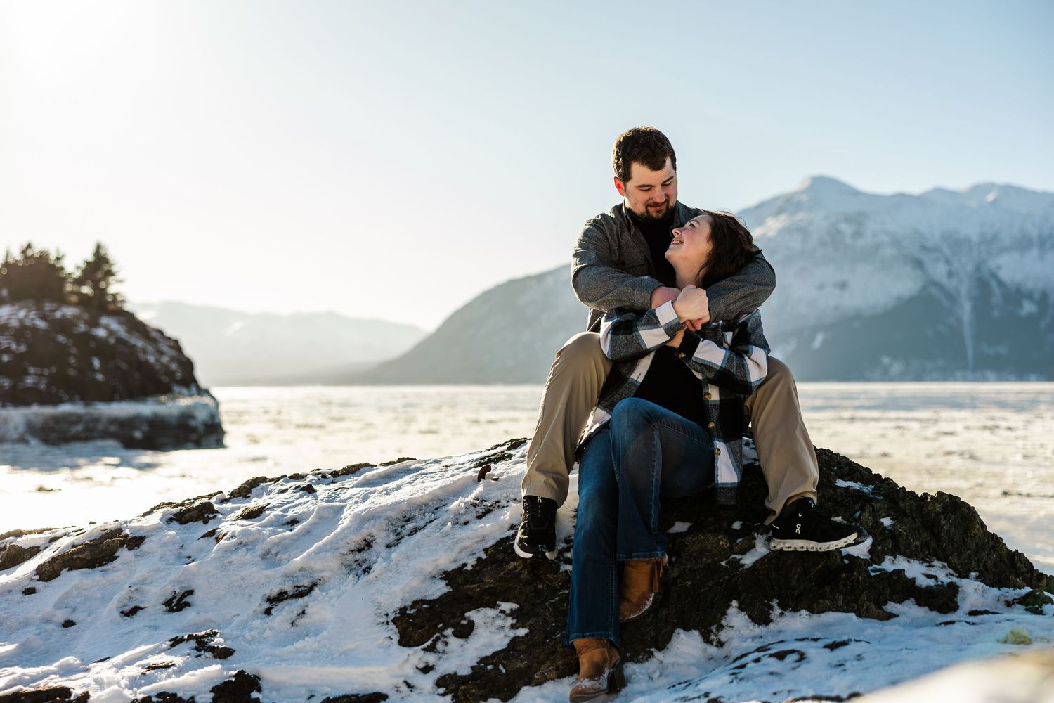 Romantic couple sitting on snowy rocks embracing with stunning winter mountain lake backdrop.