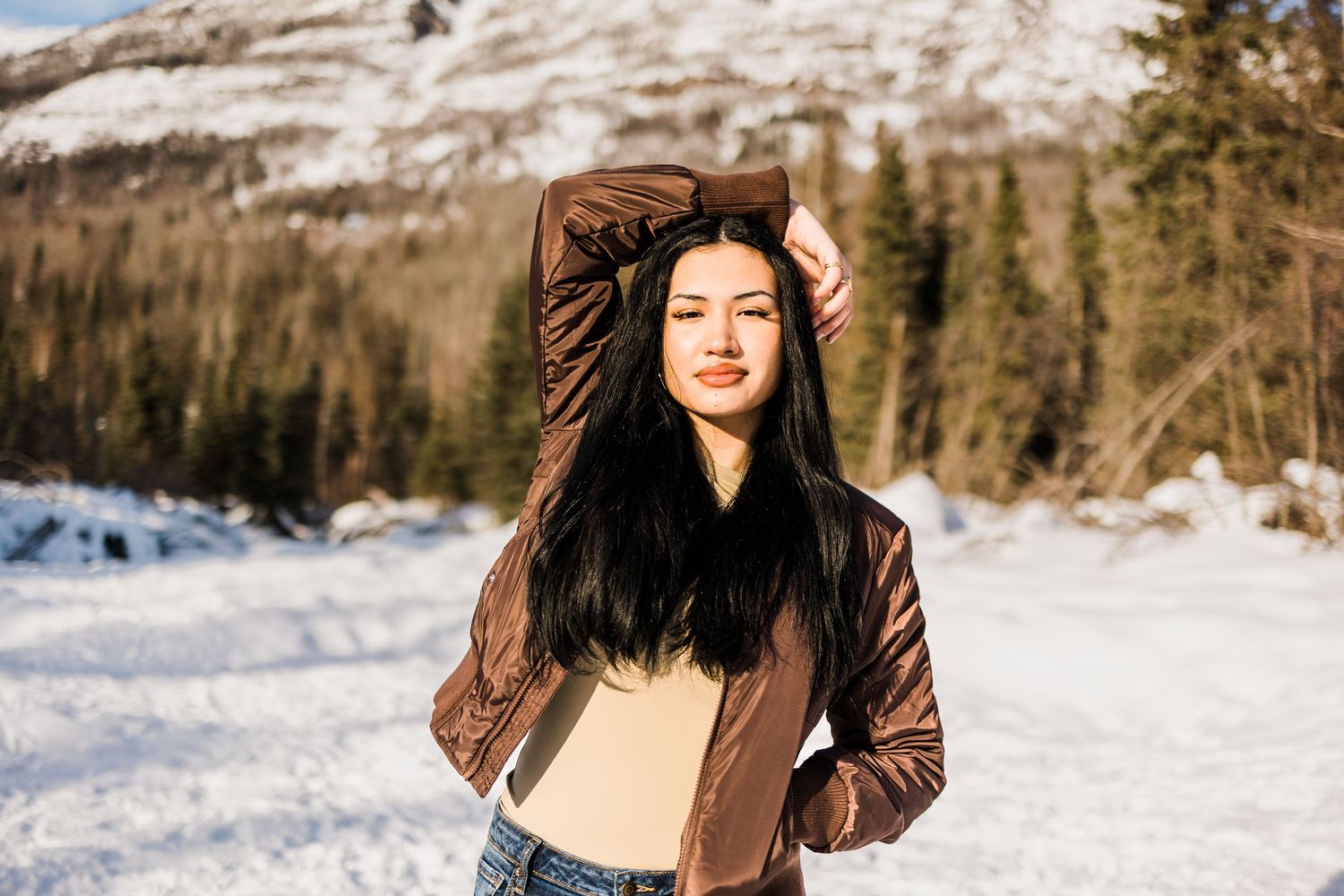 Young woman with long dark hair poses in a brown jacket against a snowy mountain landscape.