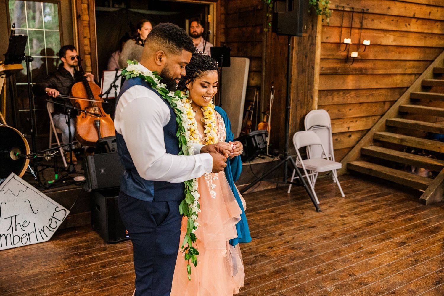 A couple in wedding attire with floral leis dance together in a wooden barn-style venue with musicians in background.