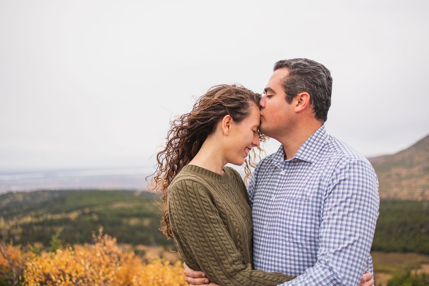 Couple sharing a romantic kiss on a hilltop with colorful autumn landscape in background.