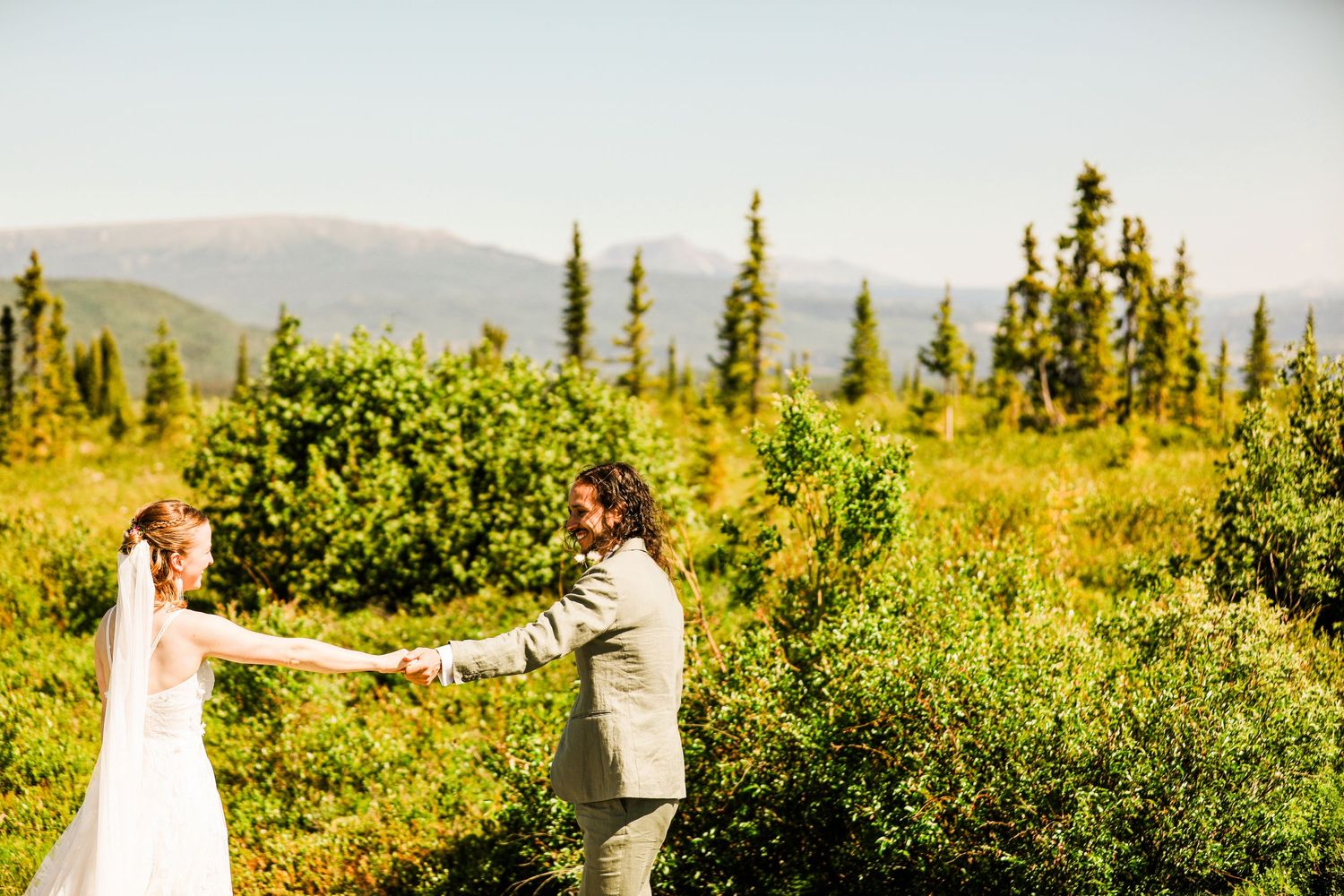 Bride and groom hold hands while dancing in mountain meadow with evergreens.