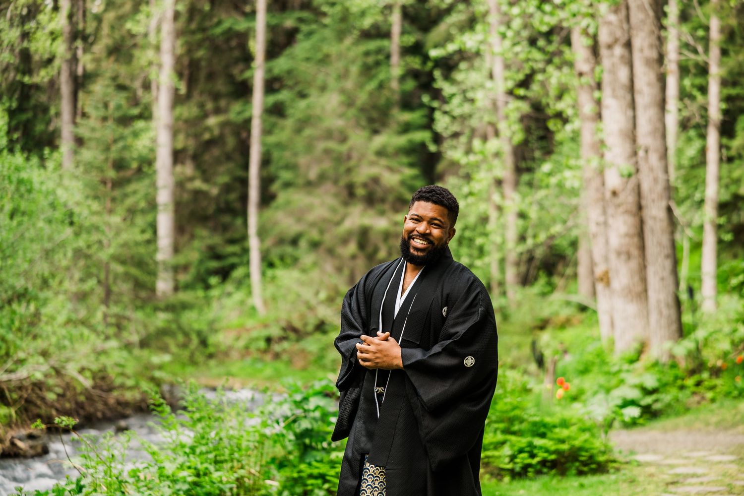 Person wearing formal black kimono stands on forest path near flowing stream.