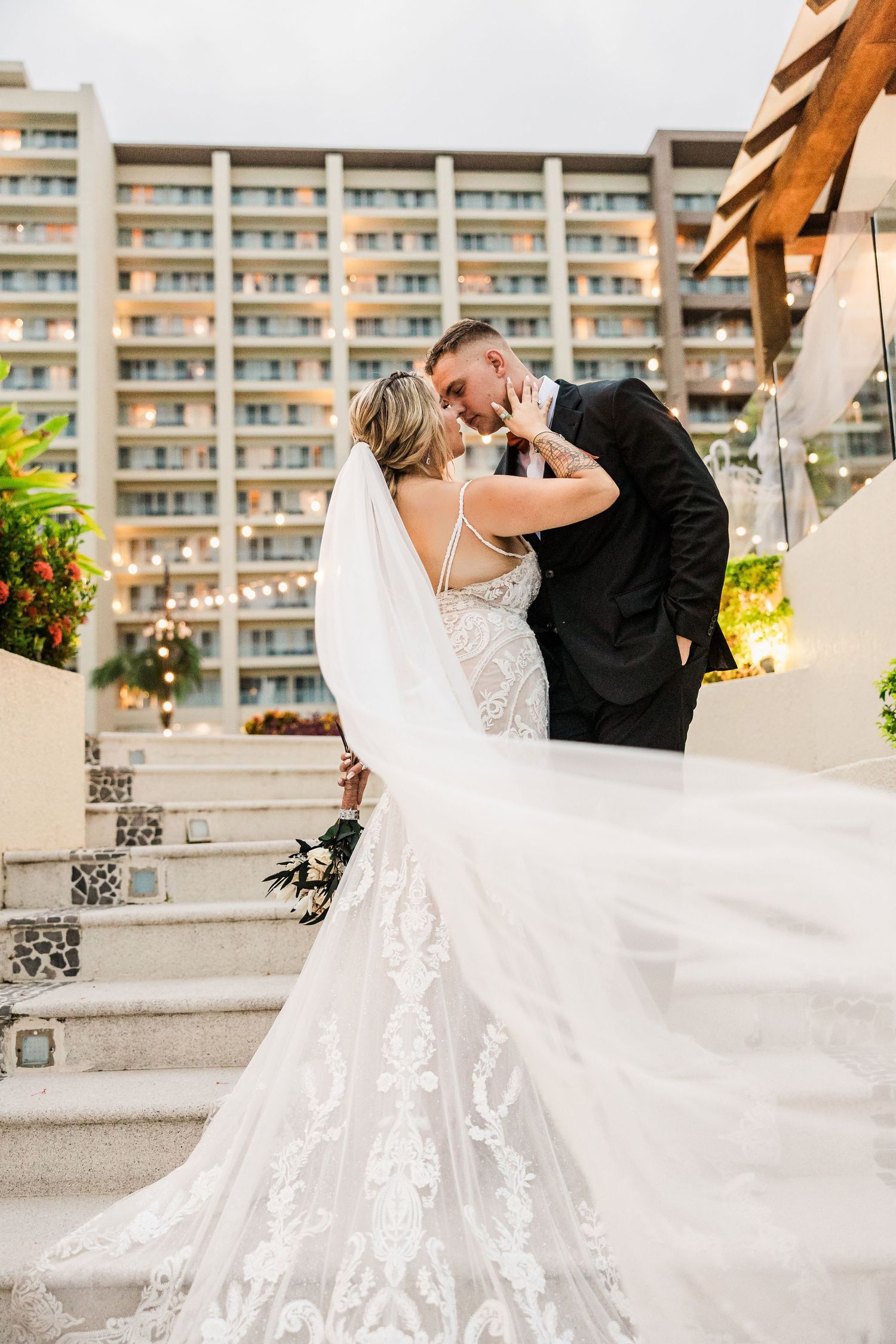 Bride and groom share a kiss on steps, her lace mermaid gown trailing, hotel building behind them.