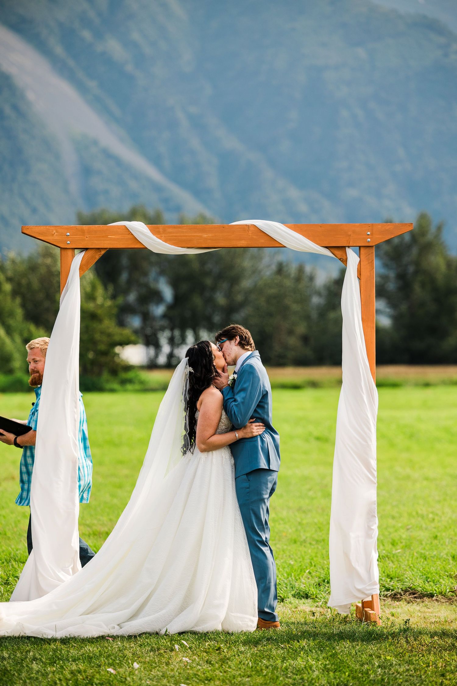 Newlyweds share intimate moment under wooden arch decorated with white fabric against mountain backdrop.