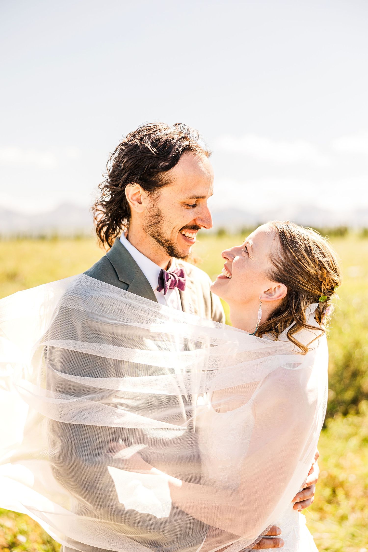 Newlywed couple embraces in golden field during sunset as bride's veil flows in the wind.