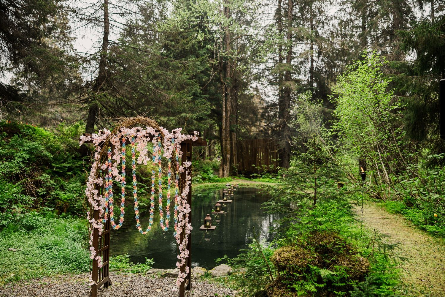 Floral wedding arch with hanging decor stands next to tranquil pond in lush forest setting.