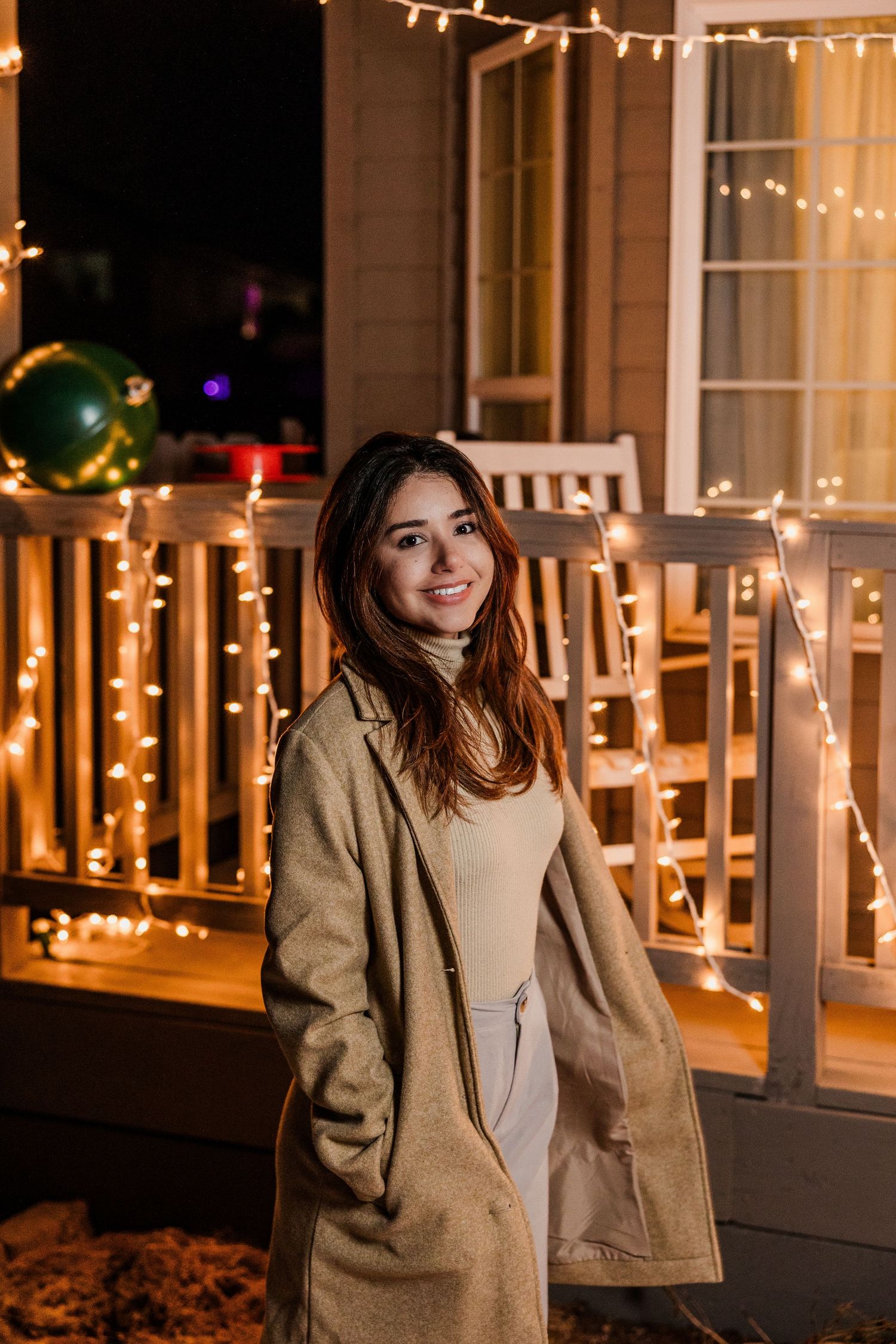 Smiling young woman in a beige coat stands on a porch decorated with warm string lights at night.