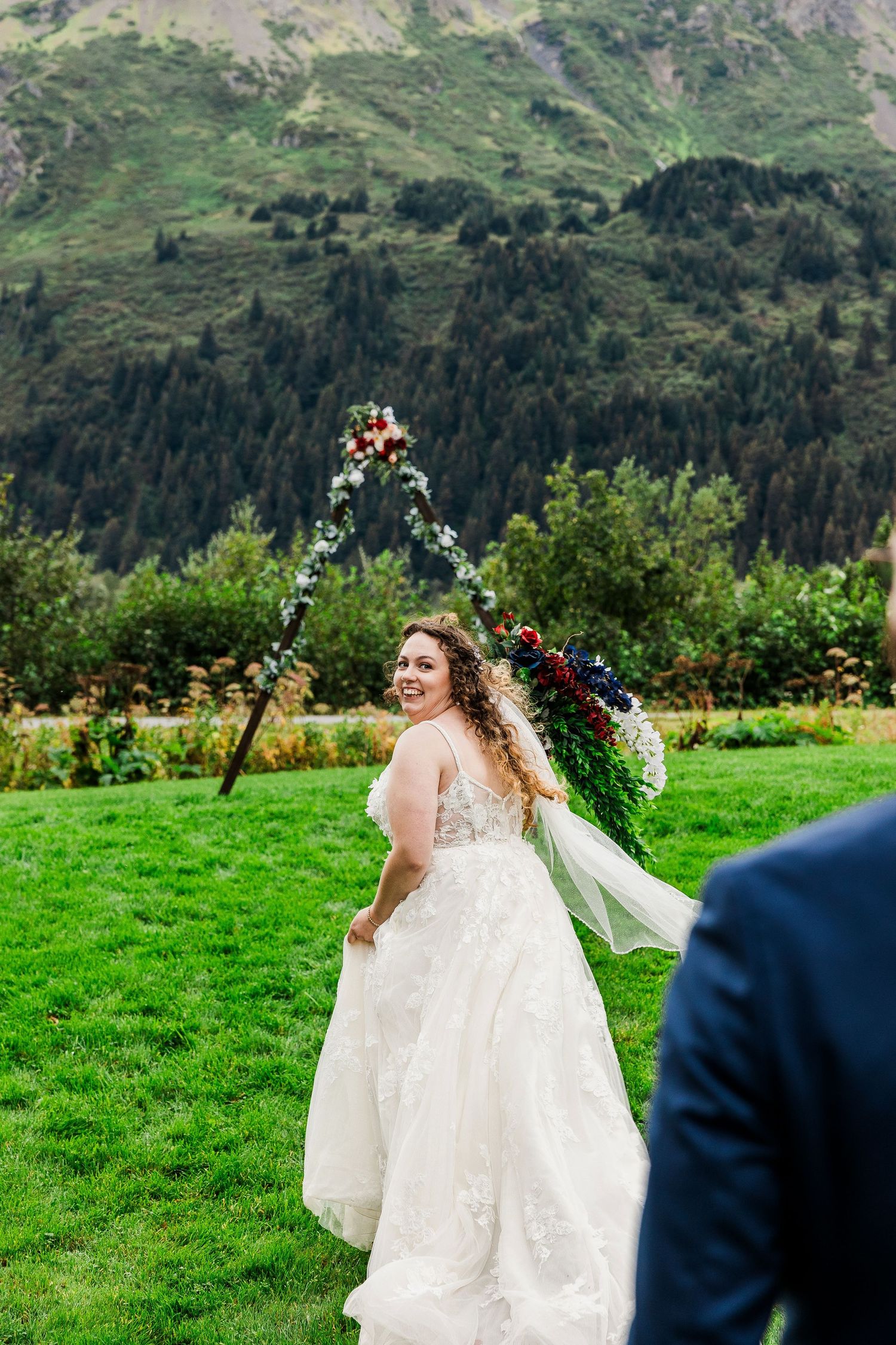 Bride in white dress twirls in mountain meadow with floral arch and stunning landscape.