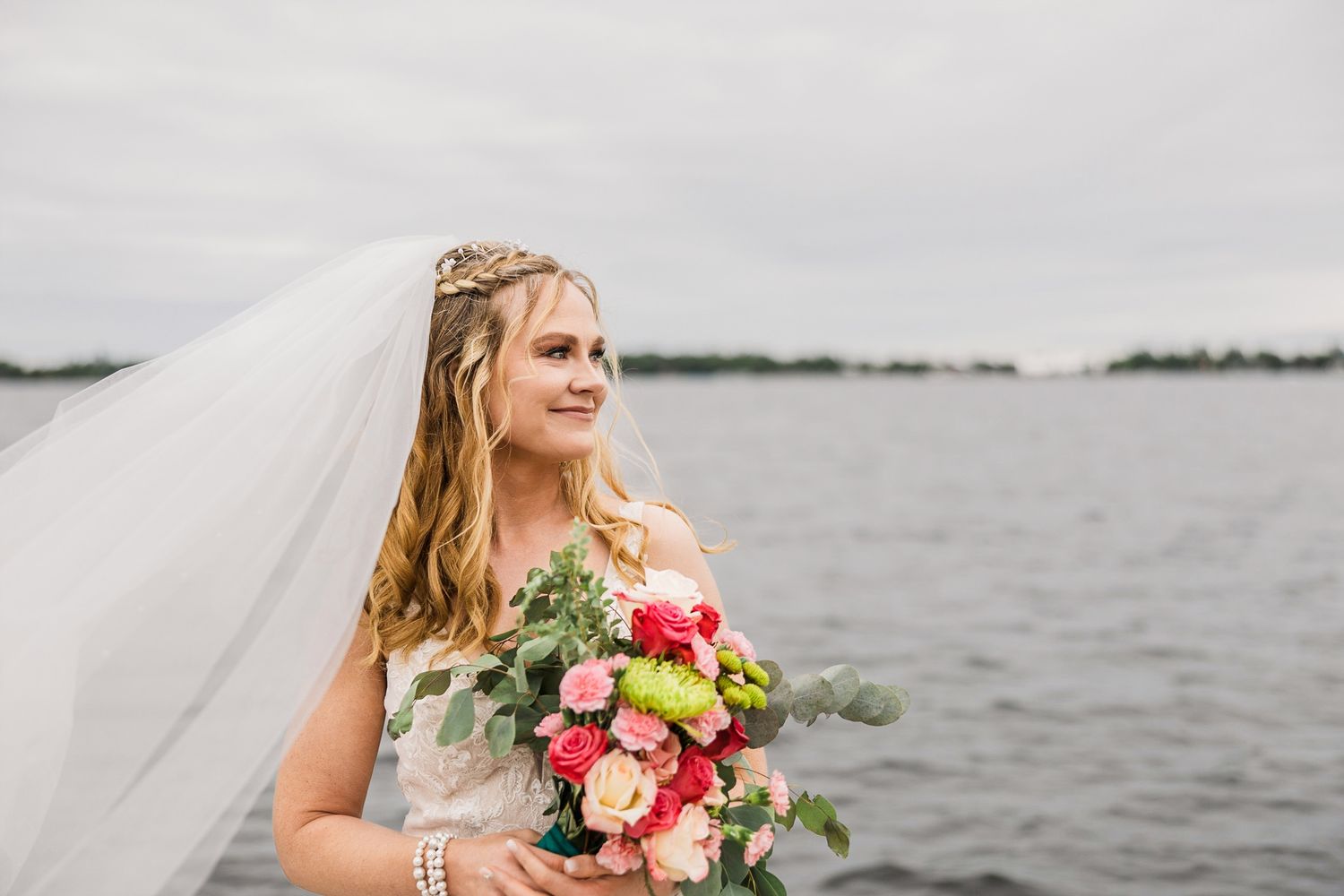 Blonde bride in white gown and veil holds bright pink and yellow bouquet beside a calm lake.