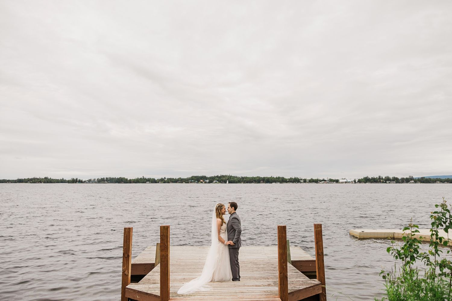 Bride and groom stand together on a wooden dock overlooking a calm lake under cloudy skies.