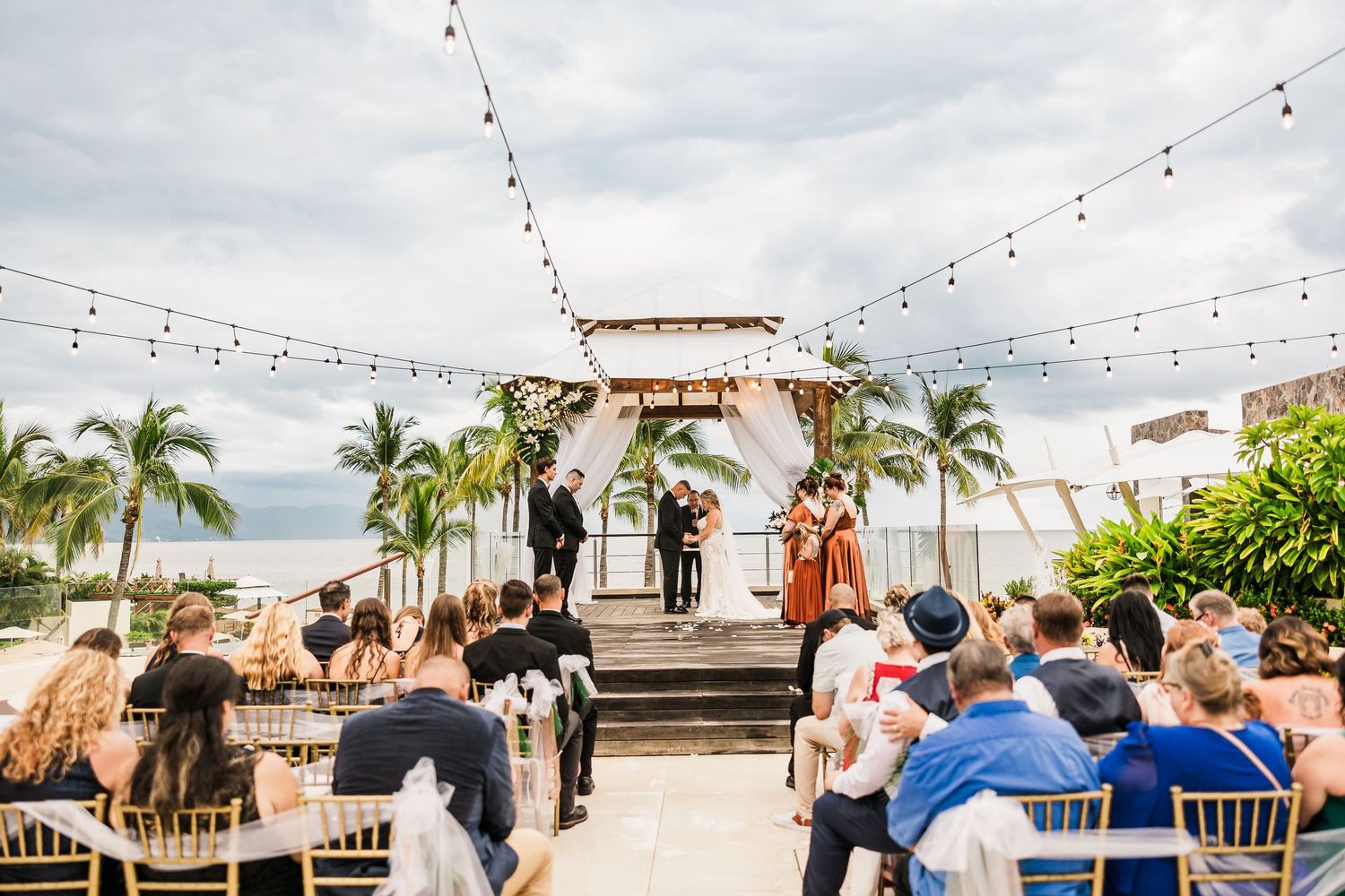 Outdoor beach wedding ceremony under string lights with palm trees and ocean backdrop.