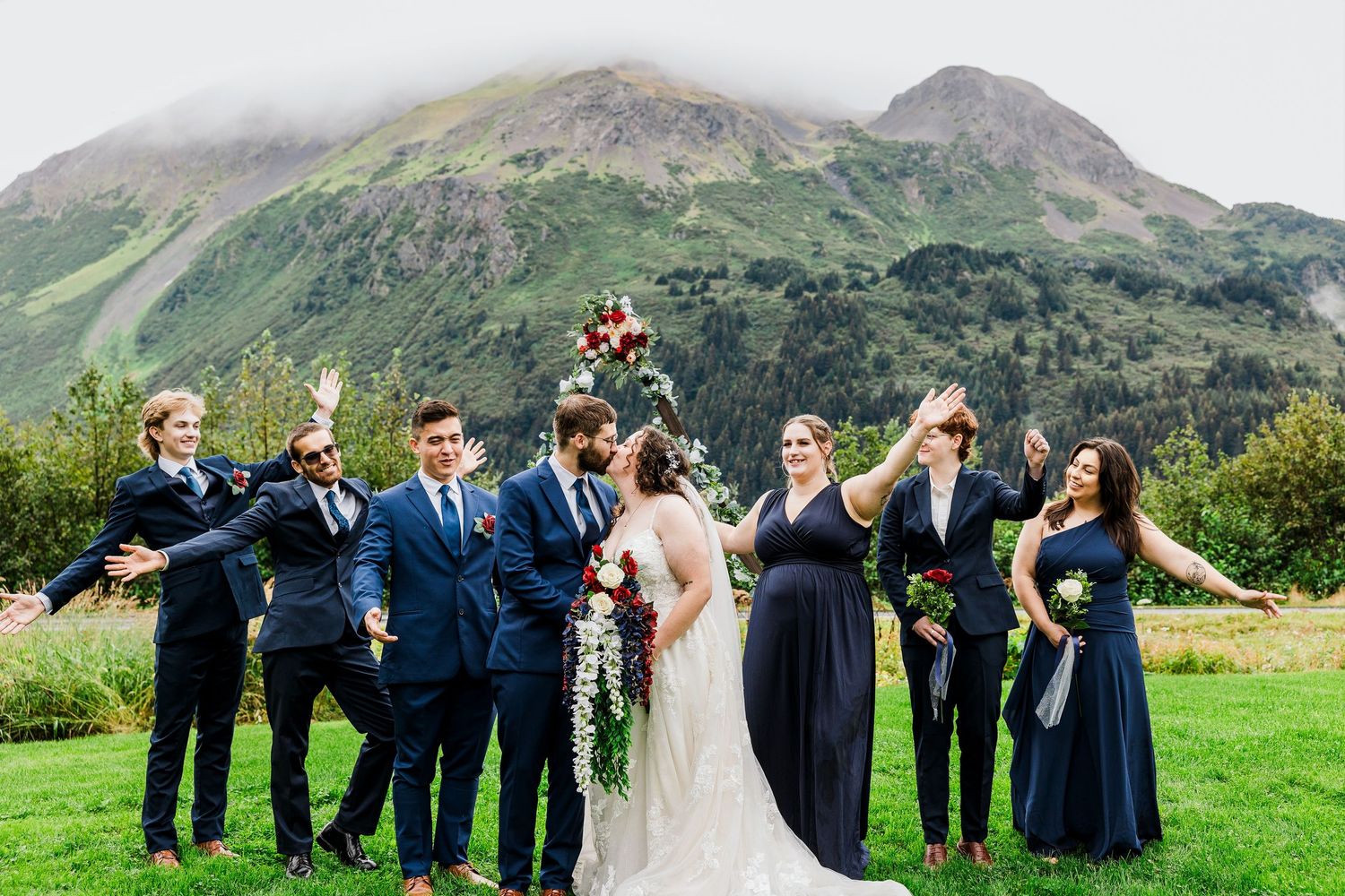 Wedding group celebrates joyfully on green lawn with majestic mountains in background.