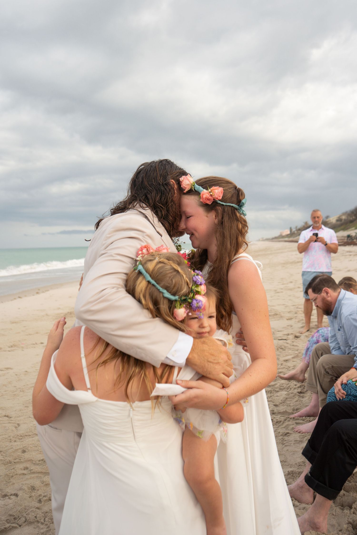 Beach wedding couple sharing a kiss while embracing children wearing floral crowns during an outdoor seaside ceremony.