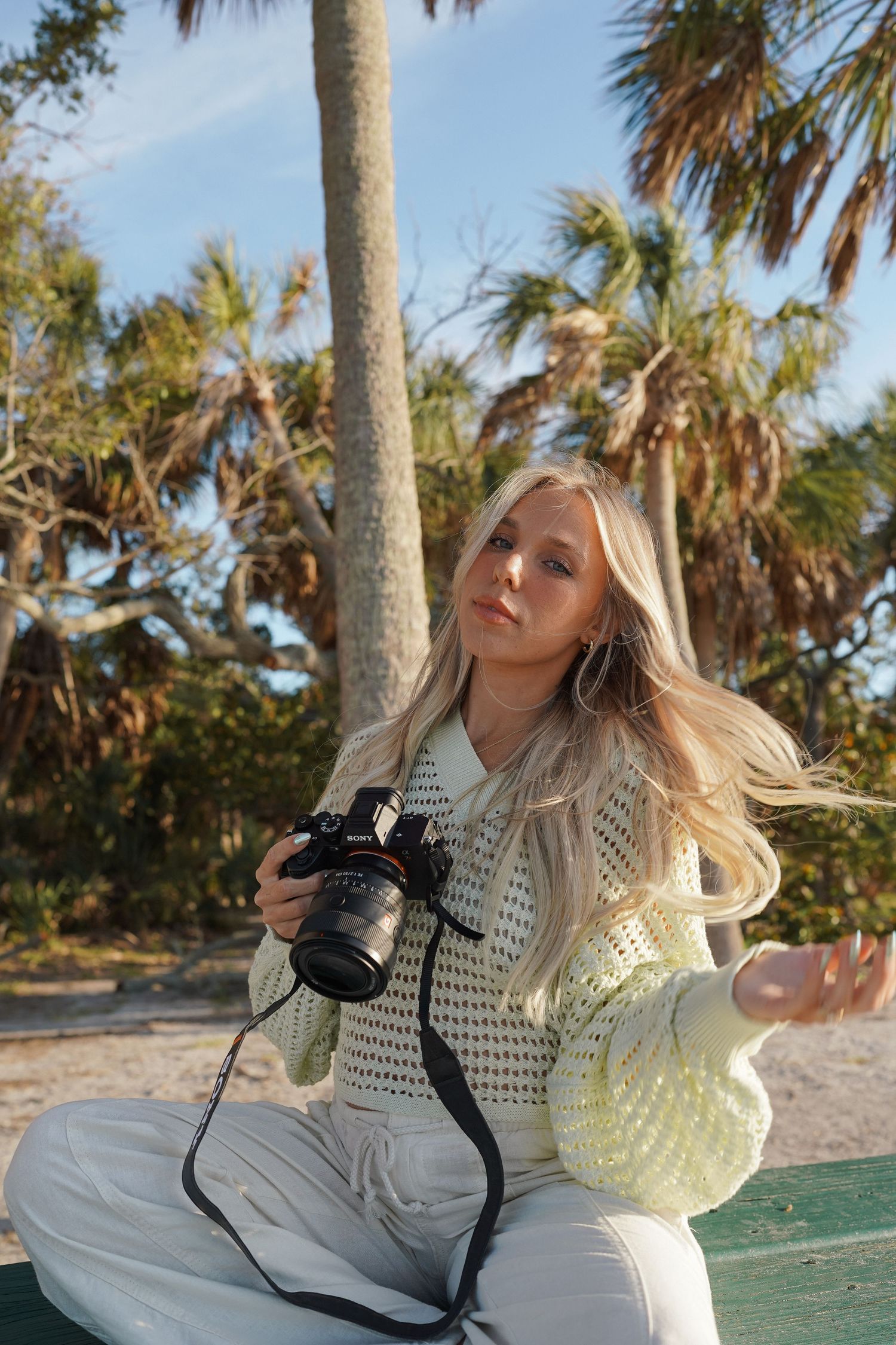 Blonde woman sitting outdoors holding a DSLR camera, wearing a white knit sweater with palm trees in the background.