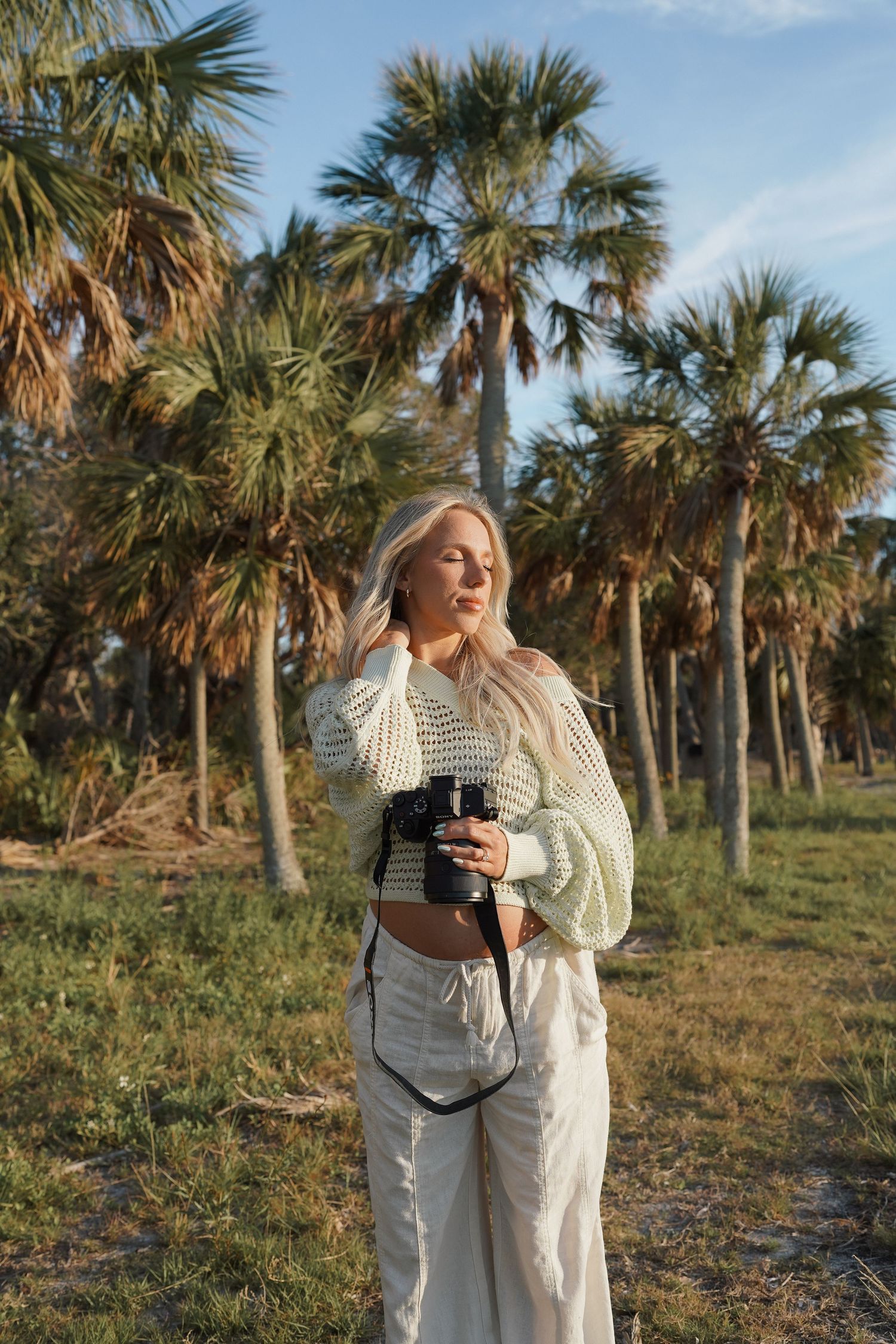 Blonde woman holding a camera in a tropical palm tree setting, wearing a white knit sweater and cream pants.