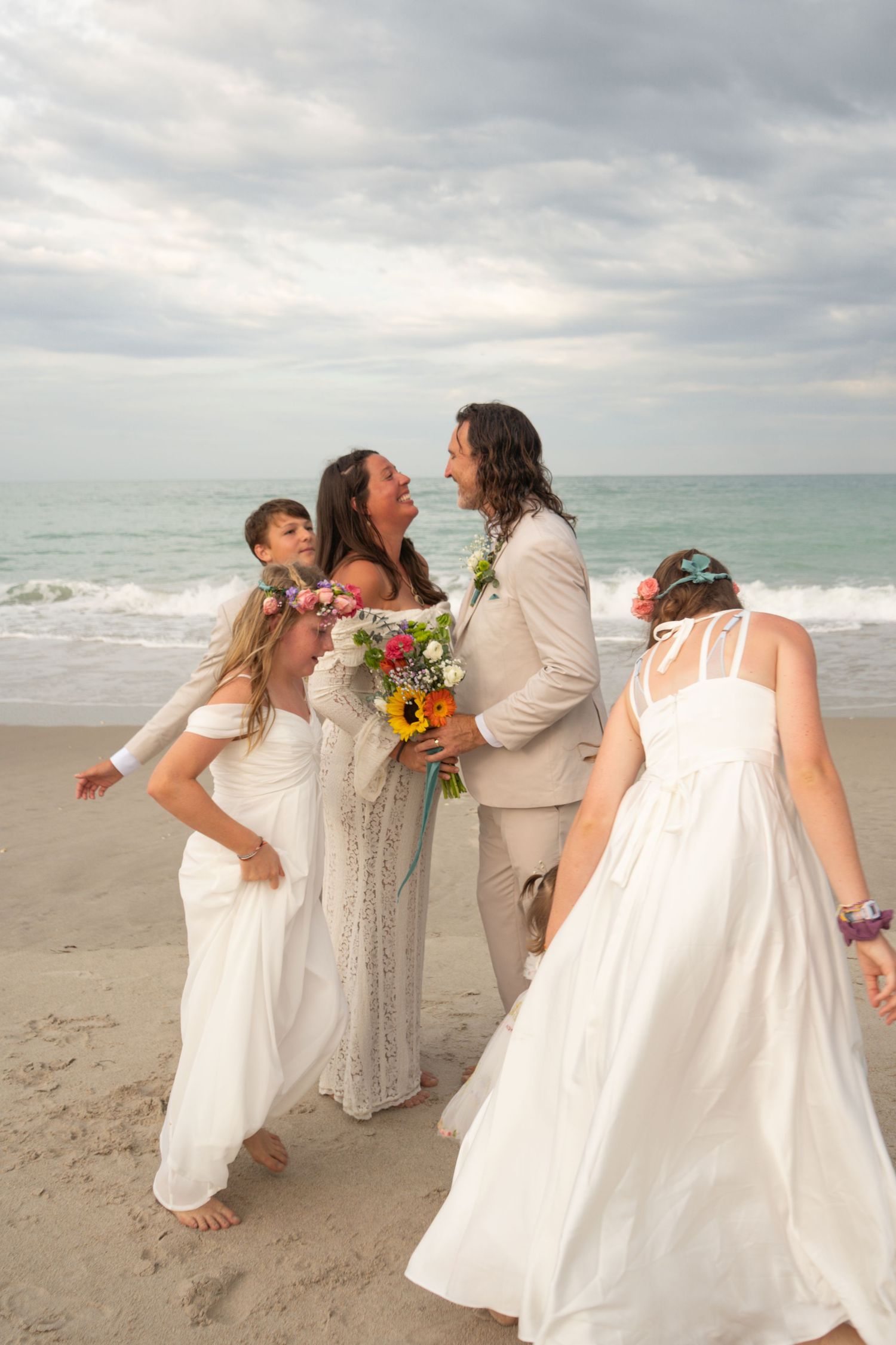 Joyful beach wedding couple in white and tan attire laughing together surrounded by children and guests on cloudy shoreline.