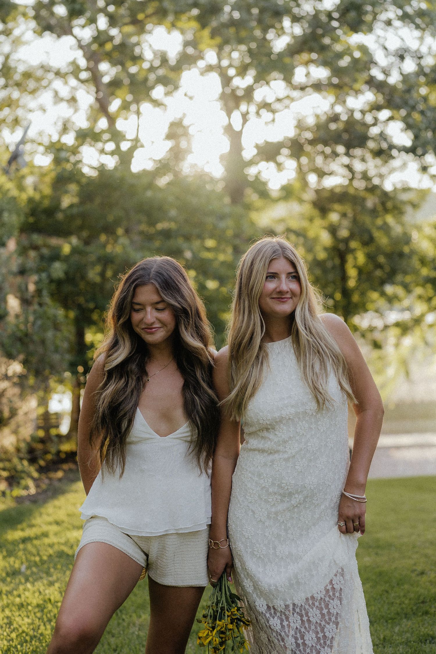 Two women in white outfits smiling together outdoors in golden sunlight with lush green trees behind.