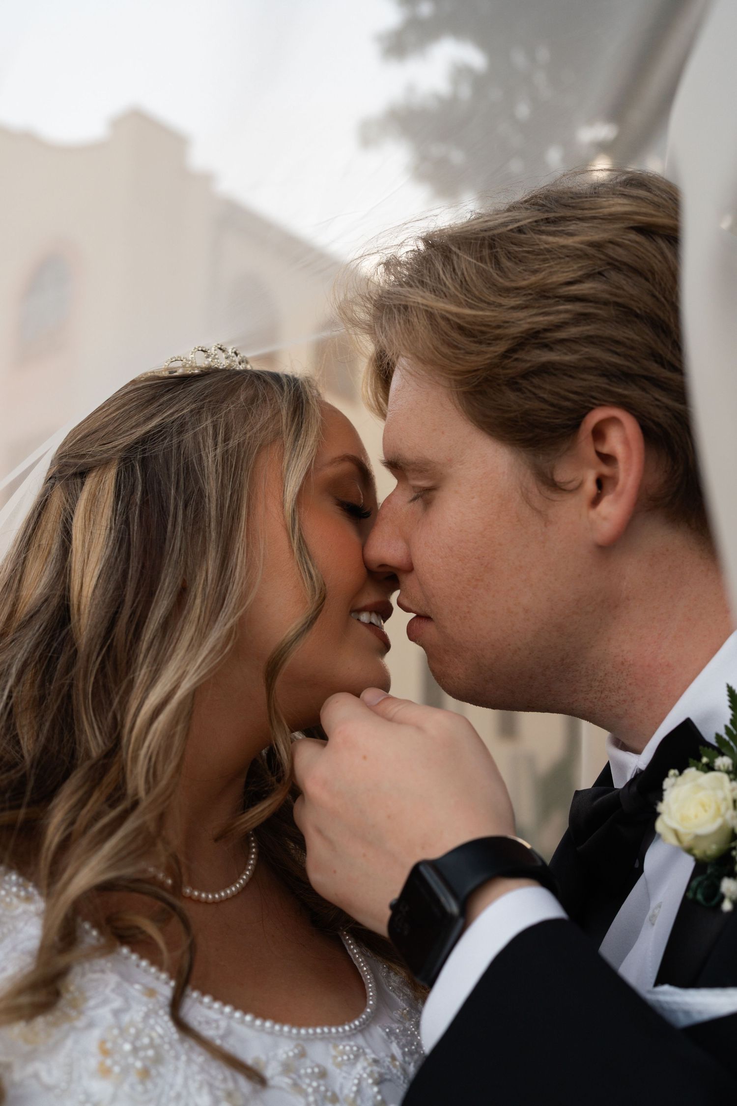 Bride and groom sharing an intimate almost-kiss moment, with groom in black tuxedo and bride wearing tiara and pearl necklace.
