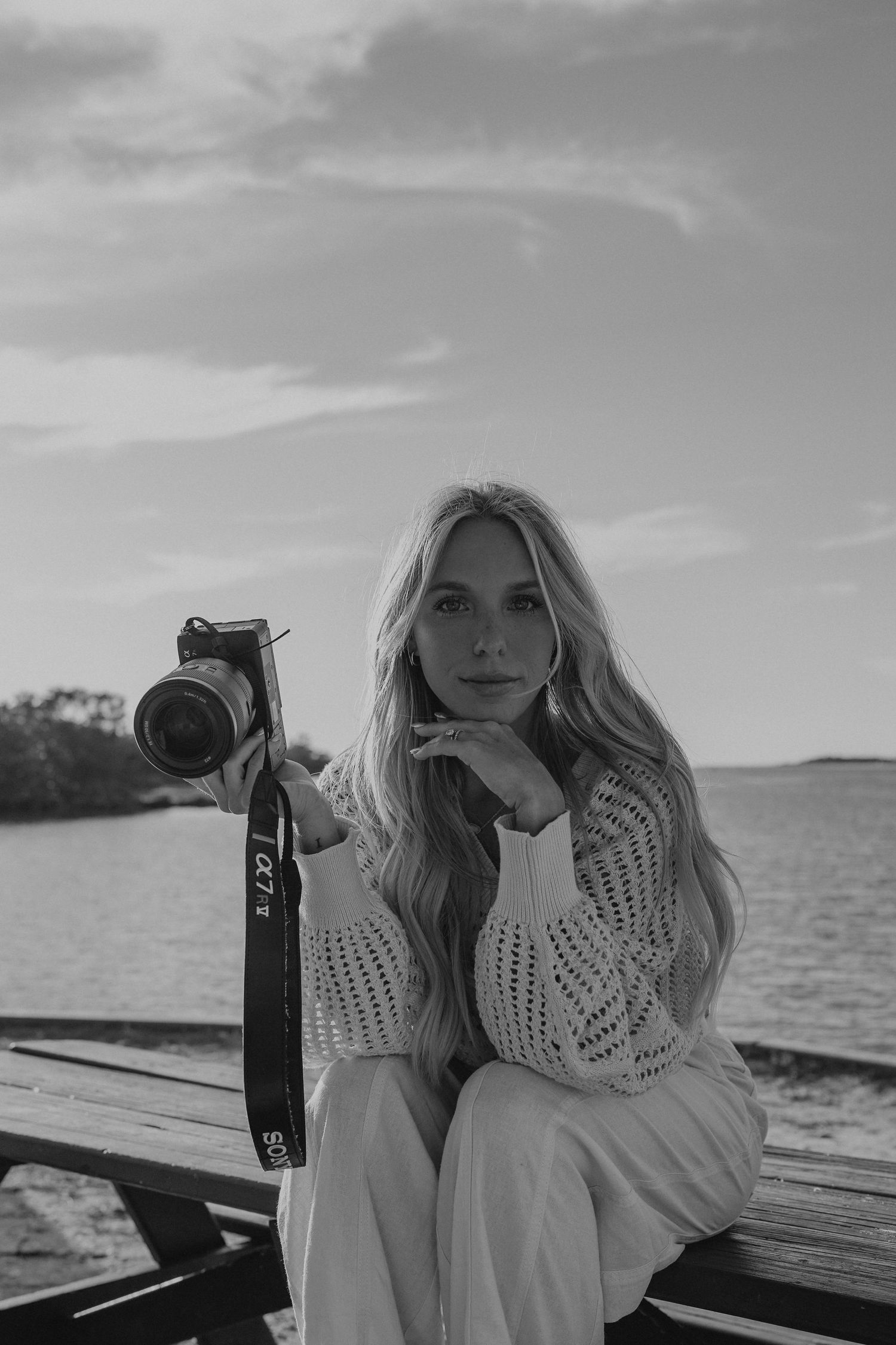 Black and white portrait of a blonde woman holding a camera, sitting on a dock by the water in a knit sweater.