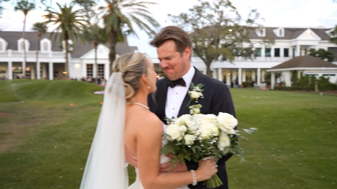 A bride and groom share a joyful moment on a lush green lawn, with the bride holding a white rose bouquet.