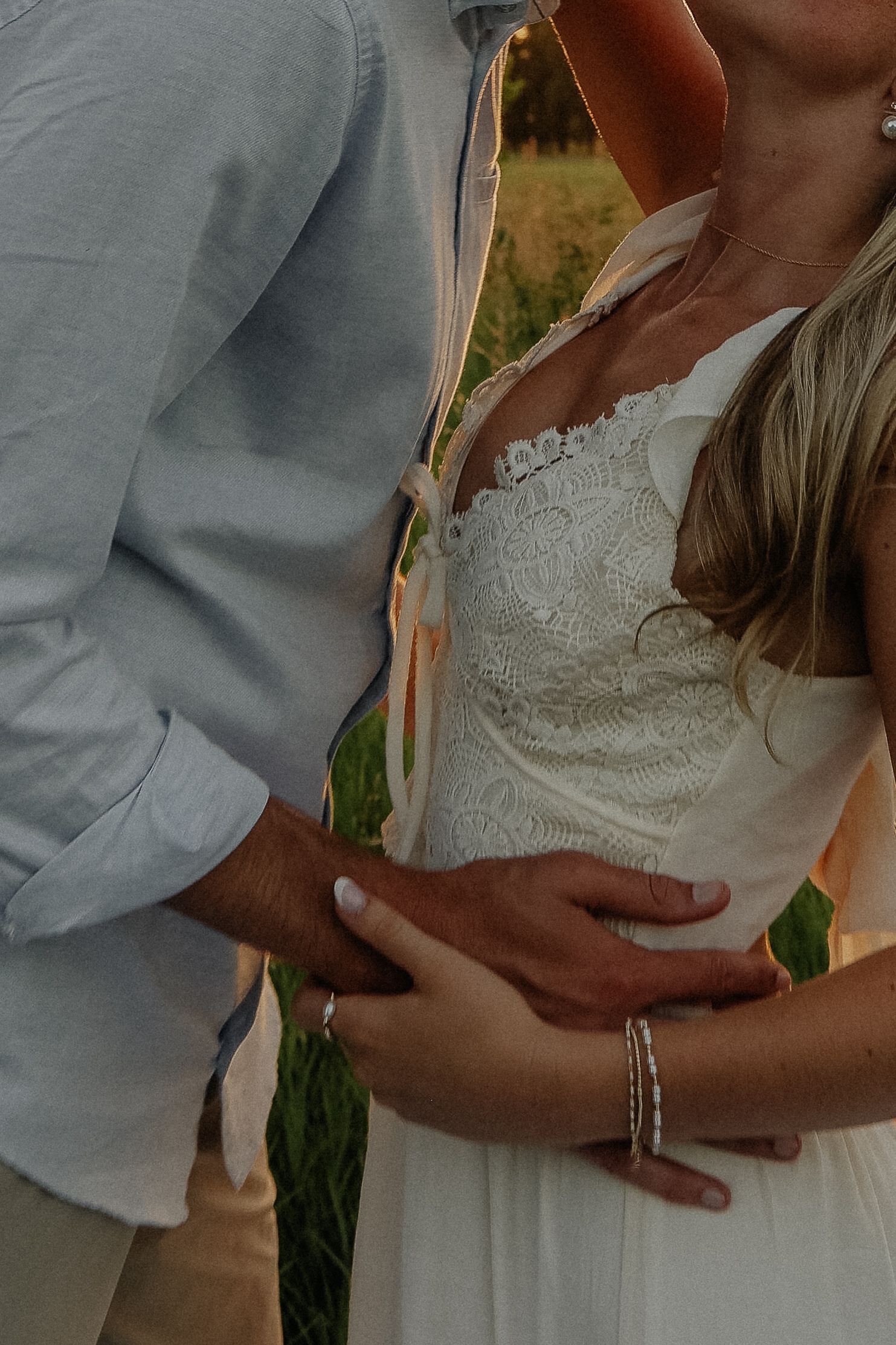 Couple tenderly embracing outdoors, woman in white lace dress with visible baby bump, man in light blue shirt holding her waist.