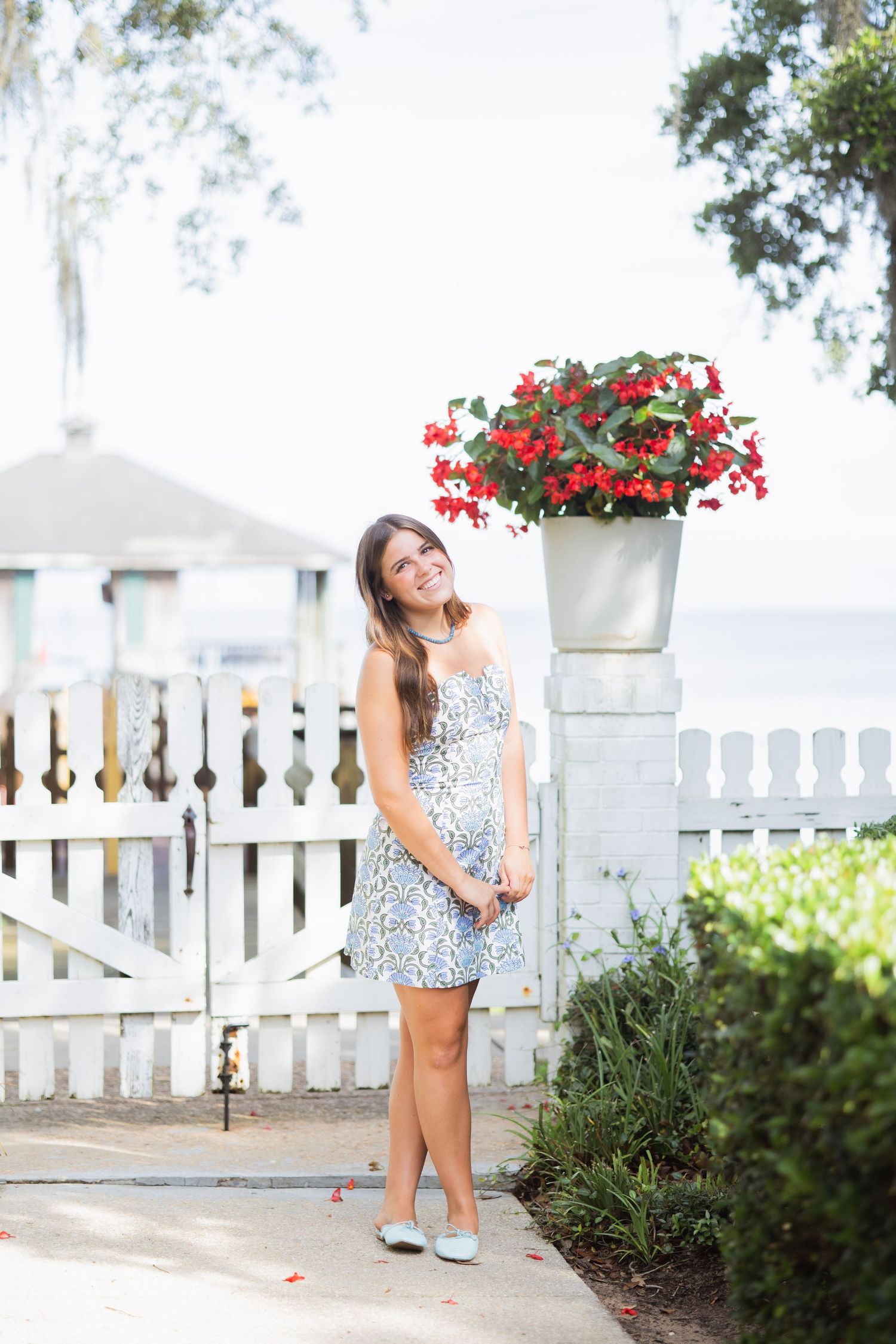 Young woman in a blue floral sundress smiling near a white picket fence with red flowers.