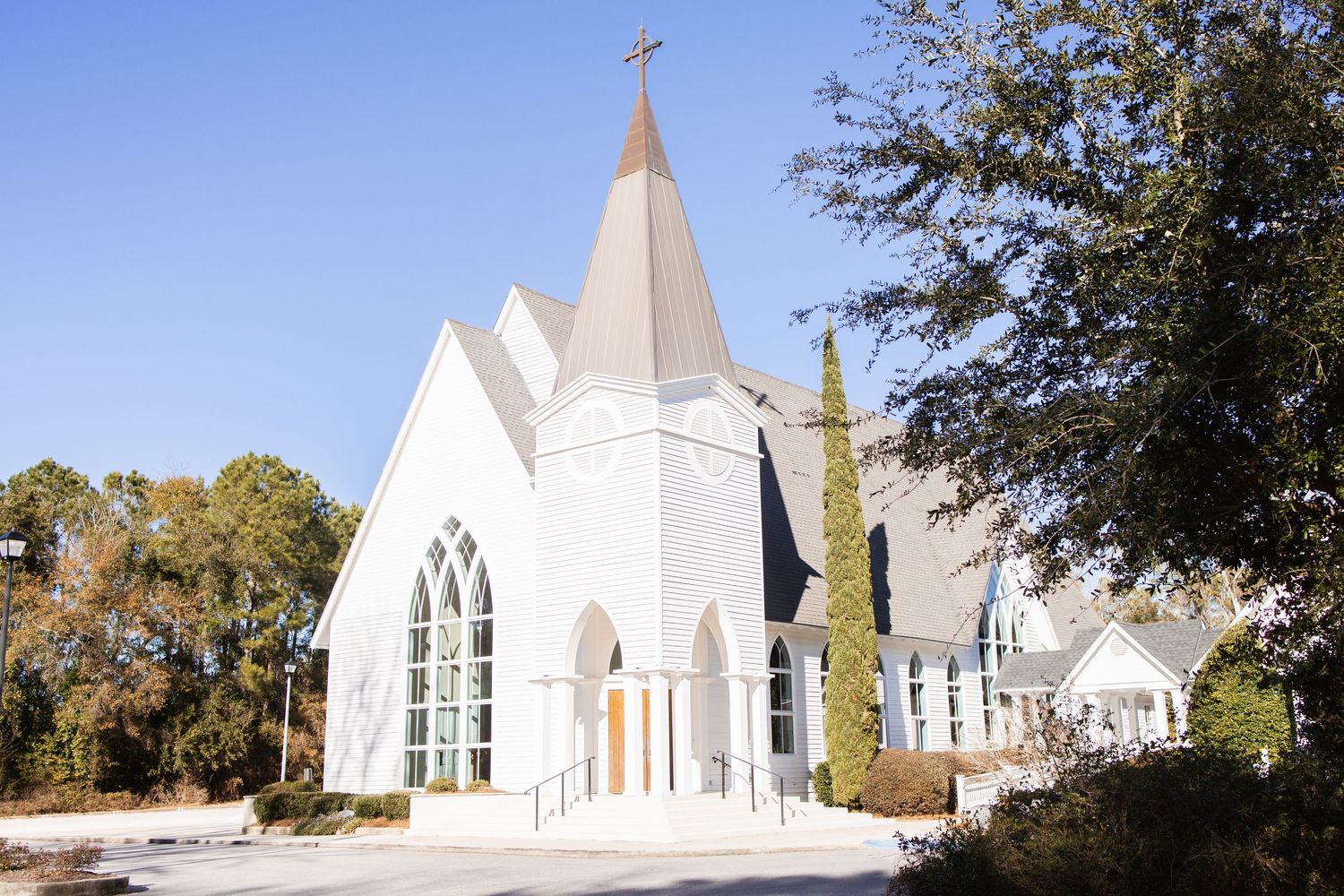 Classic white Southern church with pointed steeple and cross surrounded by tall cypress trees.