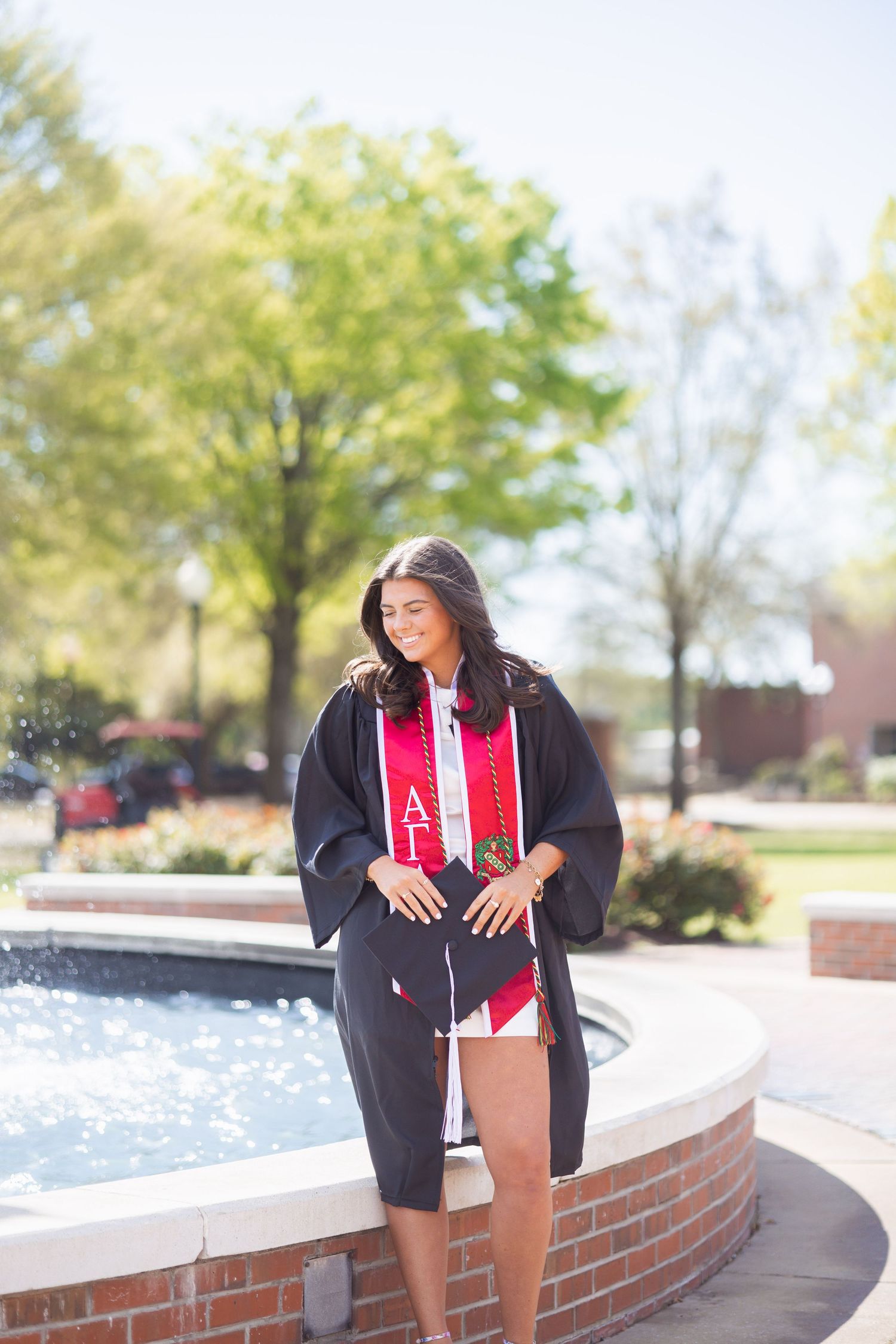Young woman in black graduation gown and red stole posing by a brick fountain on a sunny college campus.