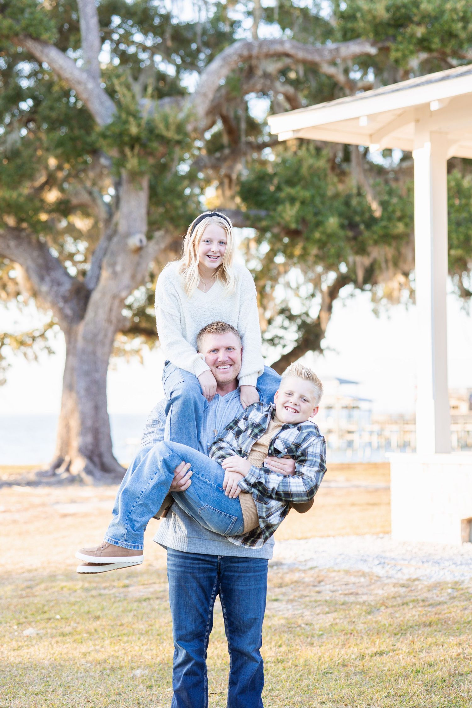 A smiling father carries his two children outdoors, with a girl on his shoulders and a boy in his arms, near a large oak tree.