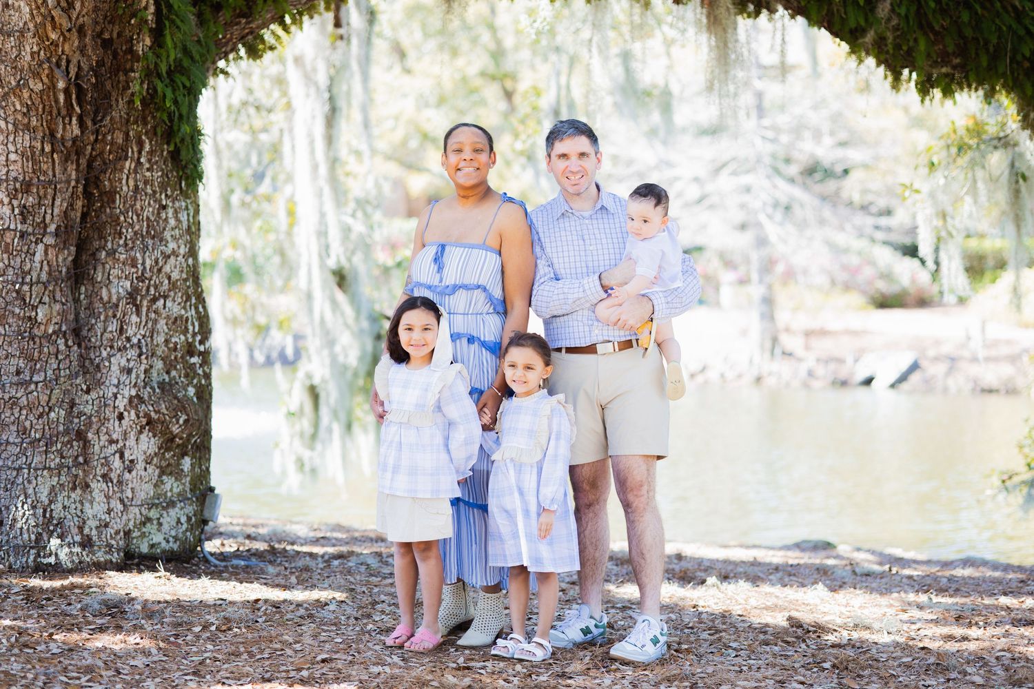 Family of five posing together outdoors near a lake under a large moss-covered tree in coordinating outfits.