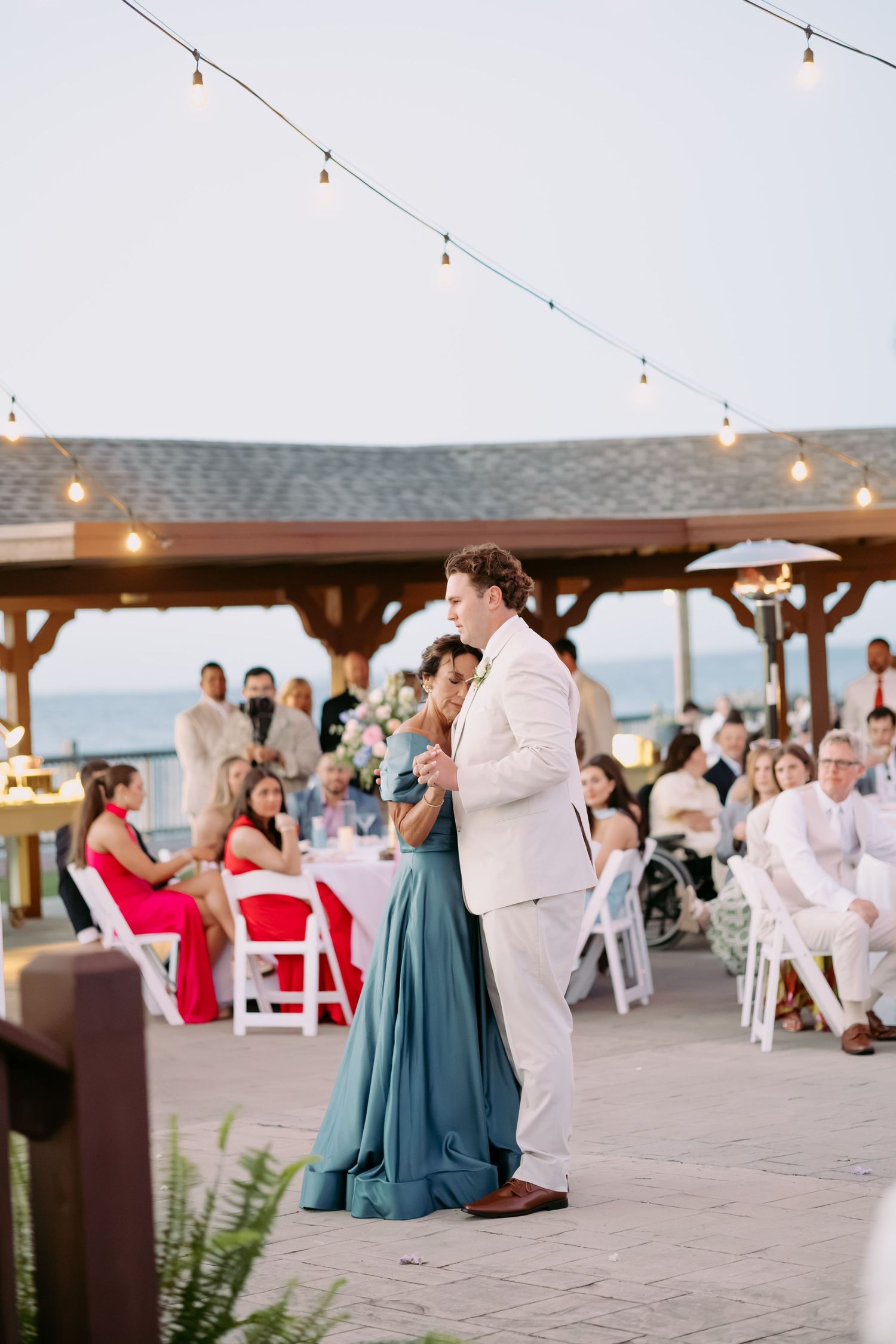 Groom in white suit shares mother-son dance at beachside outdoor wedding reception under warm string lights at dusk.