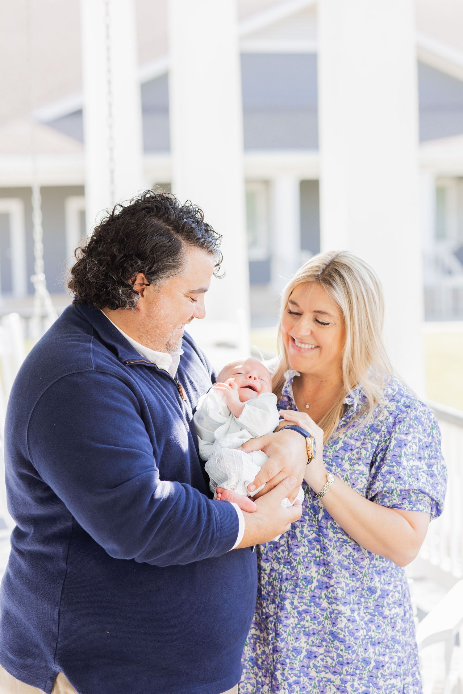 Happy parents in blue outfits lovingly gazing at their newborn baby on a bright porch.