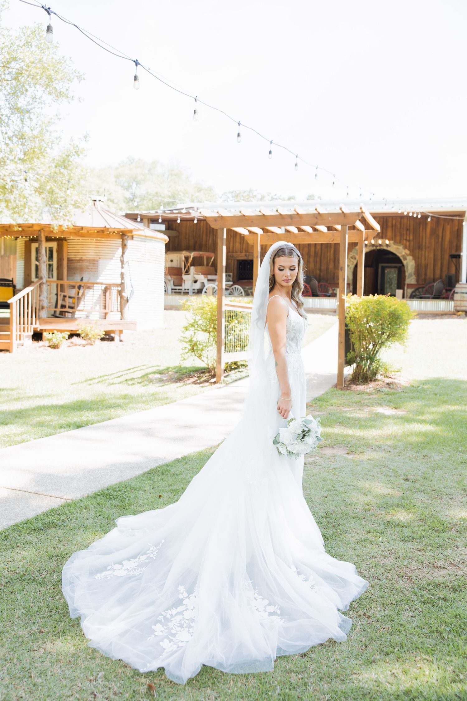 Elegant bride in lace mermaid gown with long train holds bouquet outdoors at rustic wedding venue.