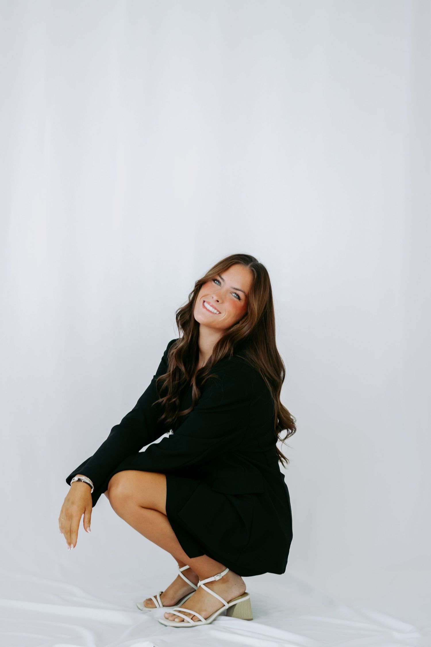 Woman in black mini dress and white strappy heels crouching and smiling against white backdrop.